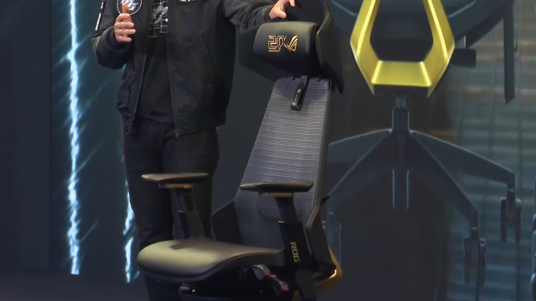A person stands next to a black and gold ASUS ROG gaming chair with '20X' on the headrest.
