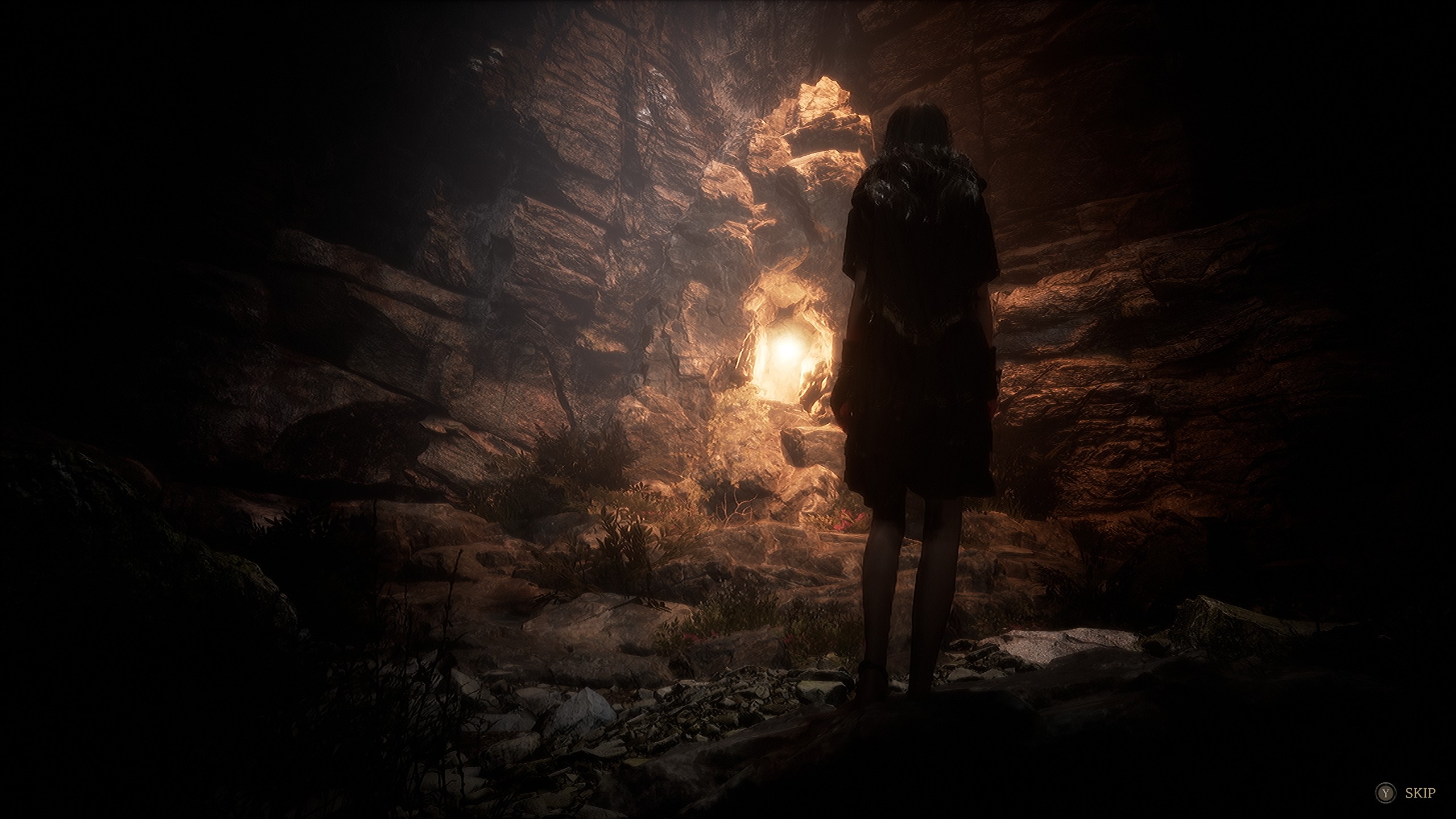A character stands in a dimly lit cave with a bright opening in the distance in a scene from a video game, with the word 'SKIP' visible in the corner.