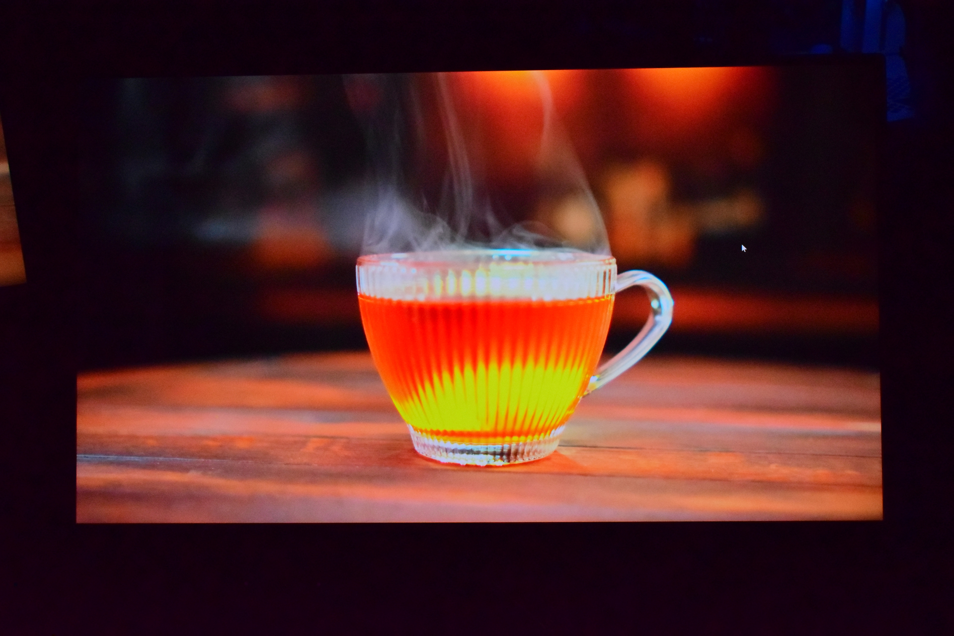 A steaming cup of tea in a transparent glass cup sits on a wooden surface with a blurred warm background.