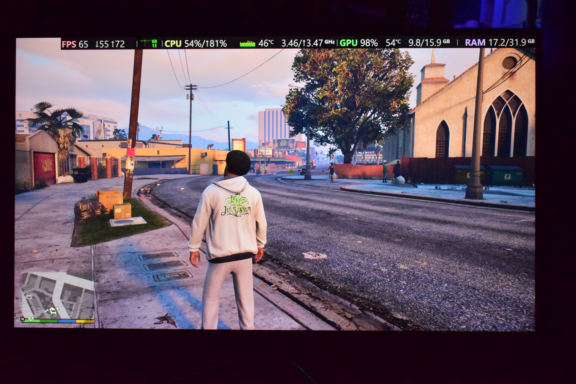 A character in 'Grand Theft Auto V' walking down a street with system metrics displayed at the top showing 'FPS 65', 'CPU 54%', and 'GPU 98%'.