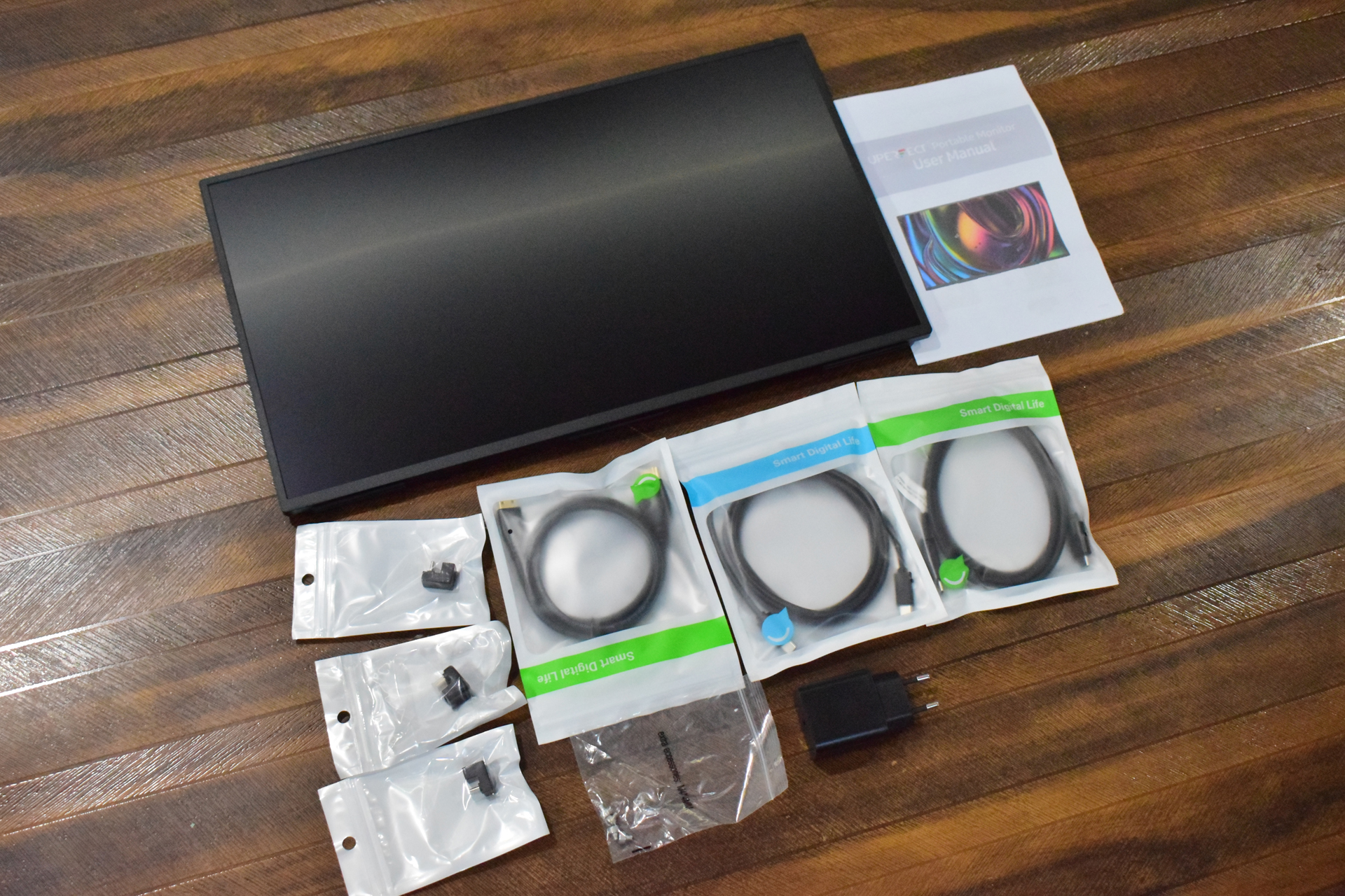 A portable monitor kit includes a black monitor, an instruction manual labeled 'User Manual,' several packaged cables from 'Smart Digital Life,' and multiple plug adapters on a wooden surface.