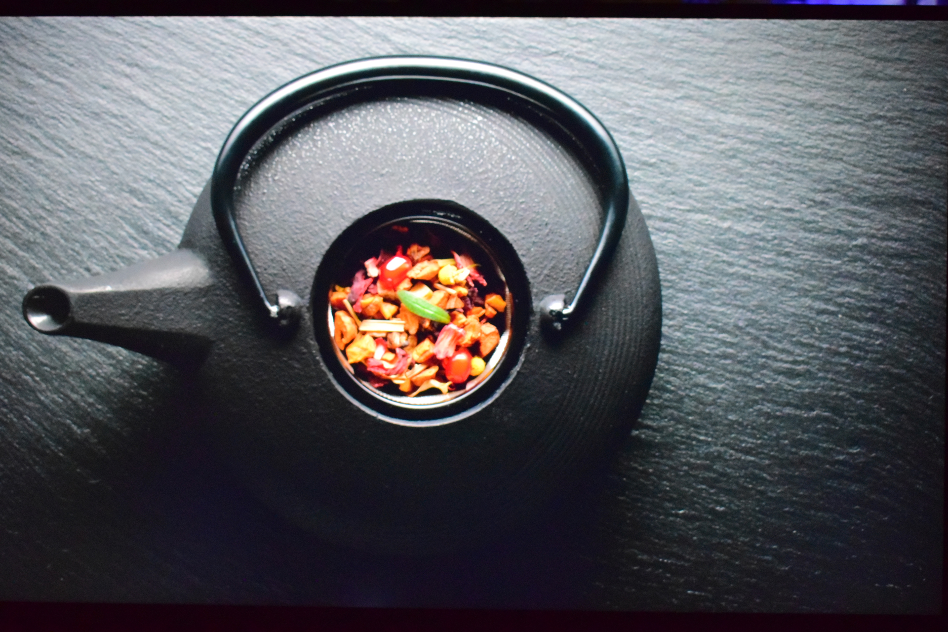 A black teapot on a slate surface with colorful dried tea leaves visible inside.