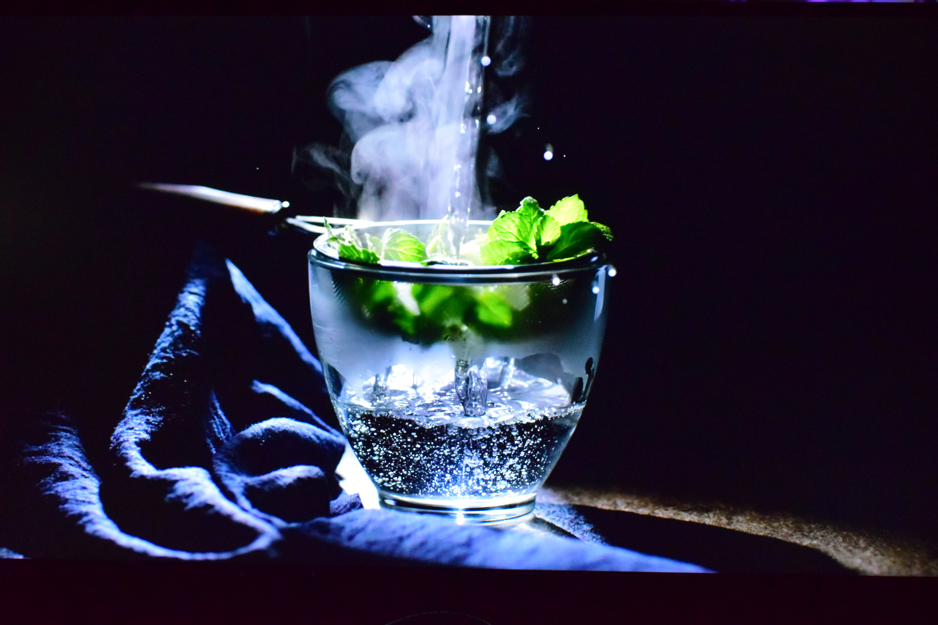 A glass cup is filled with a bubbling liquid and mint leaves, with steam rising above, set against a dark background with a blue cloth on the side.