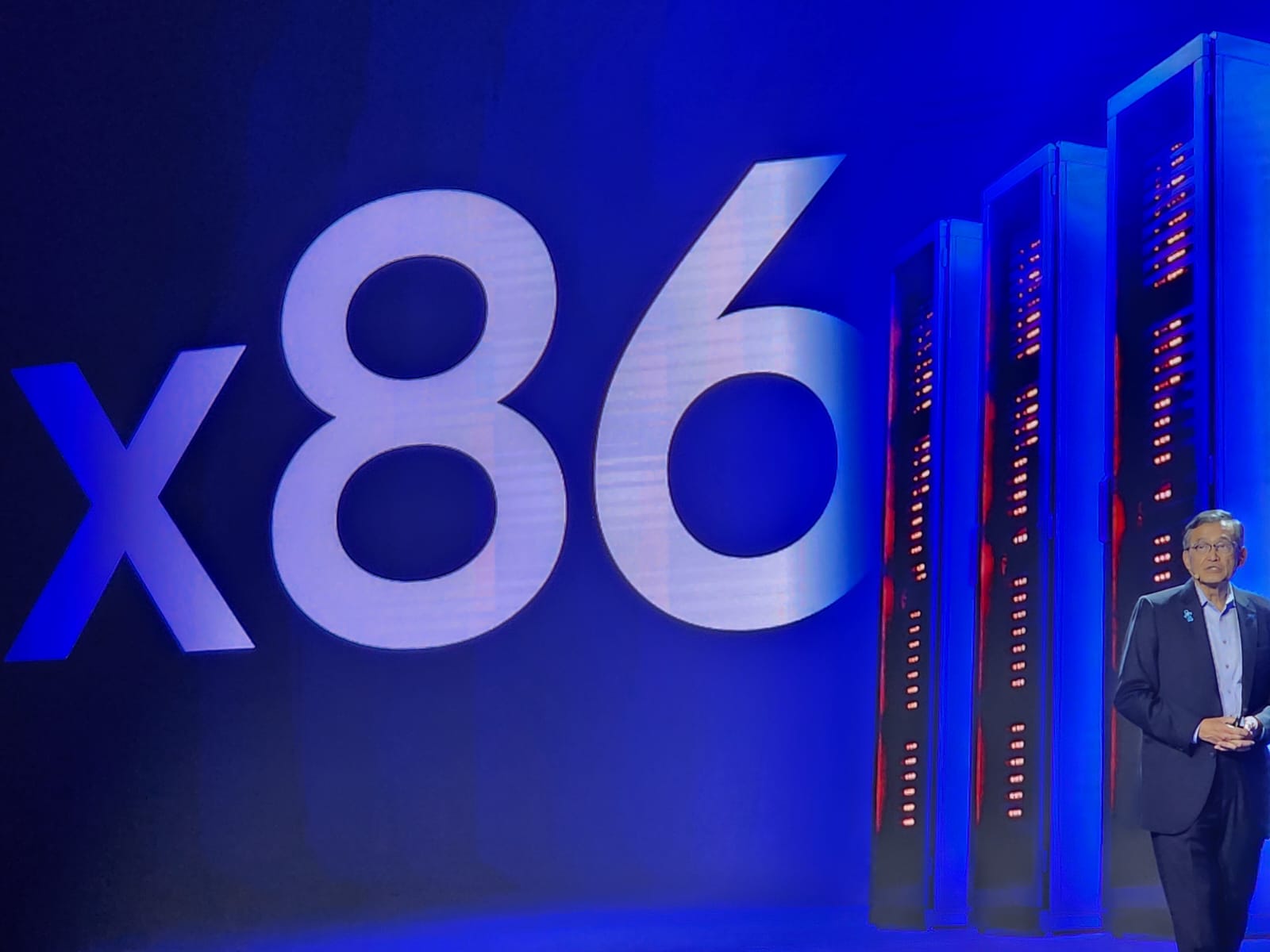 A person stands on stage next to a large 'x86' graphic and several tall server racks.