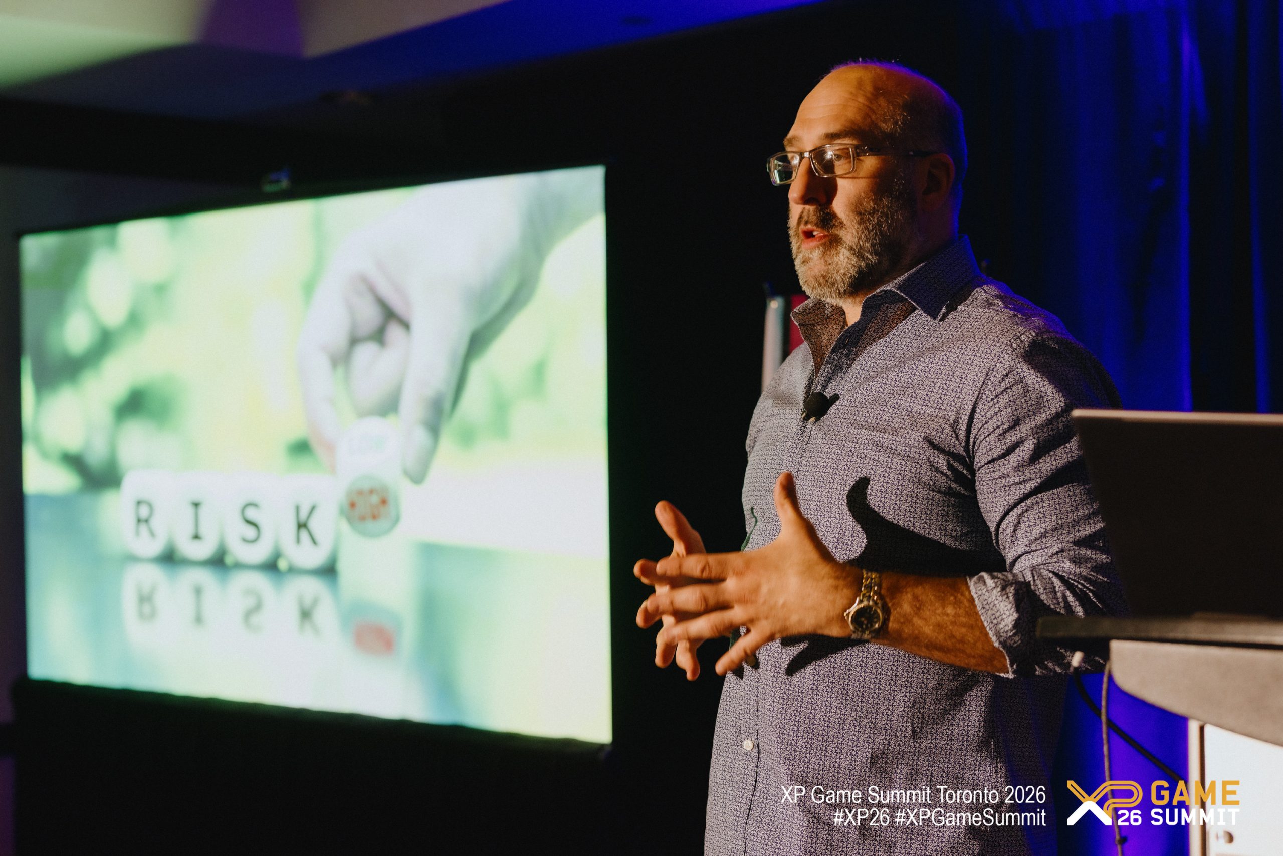 Jason Della Rocca speaking at the XP Game Summit Toronto 2026 stands next to a presentation screen displaying the word 'RISK' formed by dice.