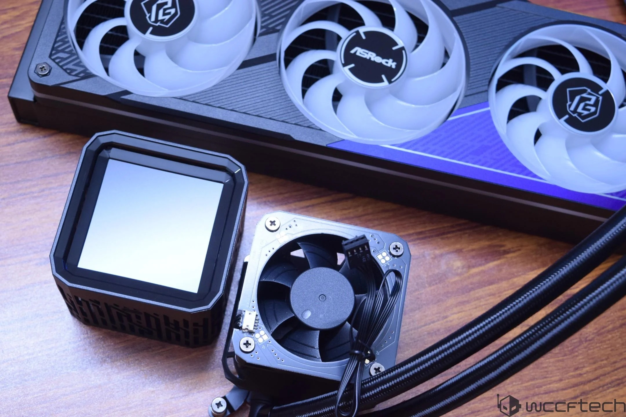 An ASRock cooling system featuring a radiator with dual fans and a pump unit with a fan and cable is displayed on a wooden surface.