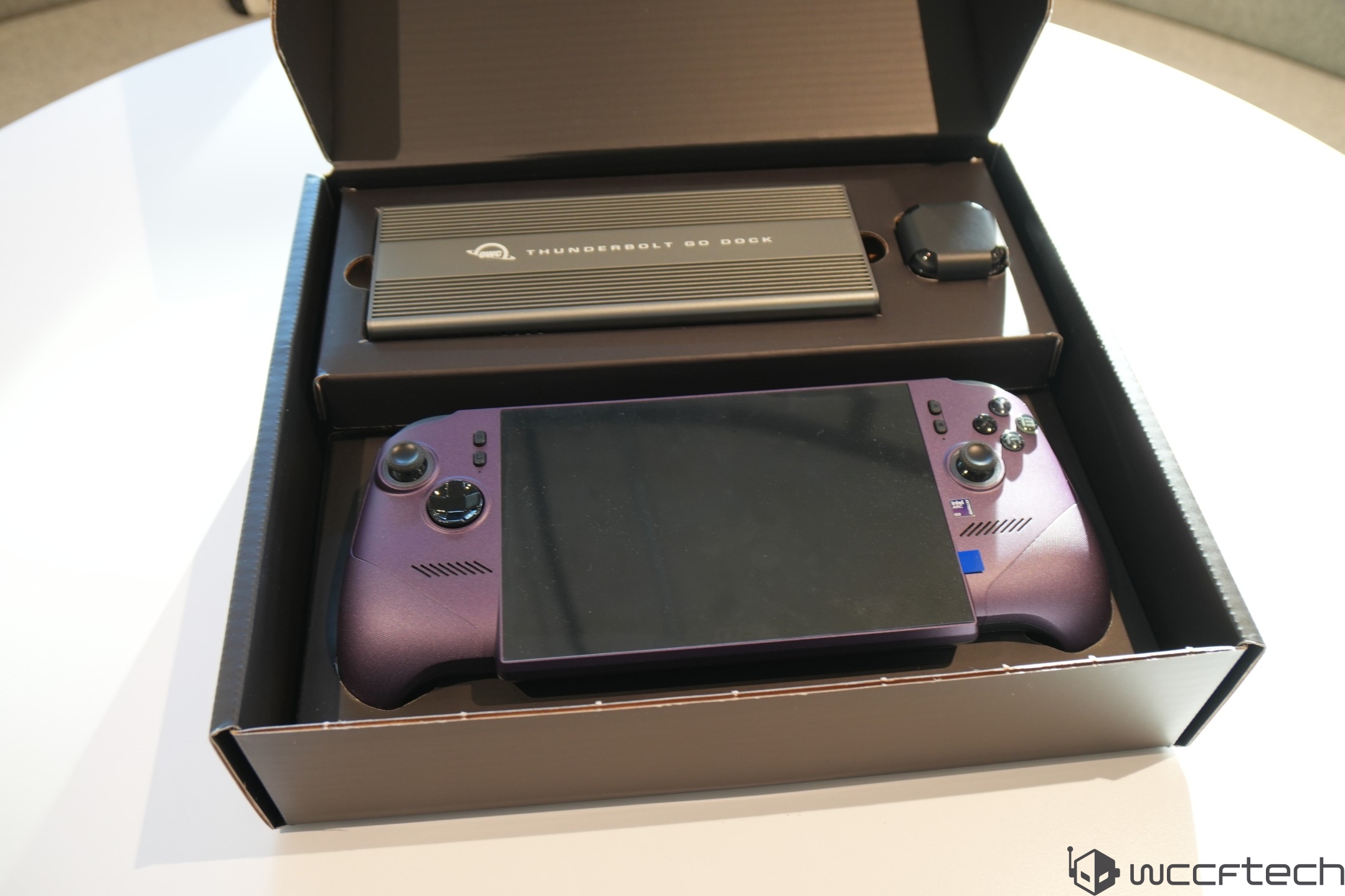 A purple handheld gaming console is packaged alongside an OWC Thunderbolt Go Dock and charging cable in a cardboard box.