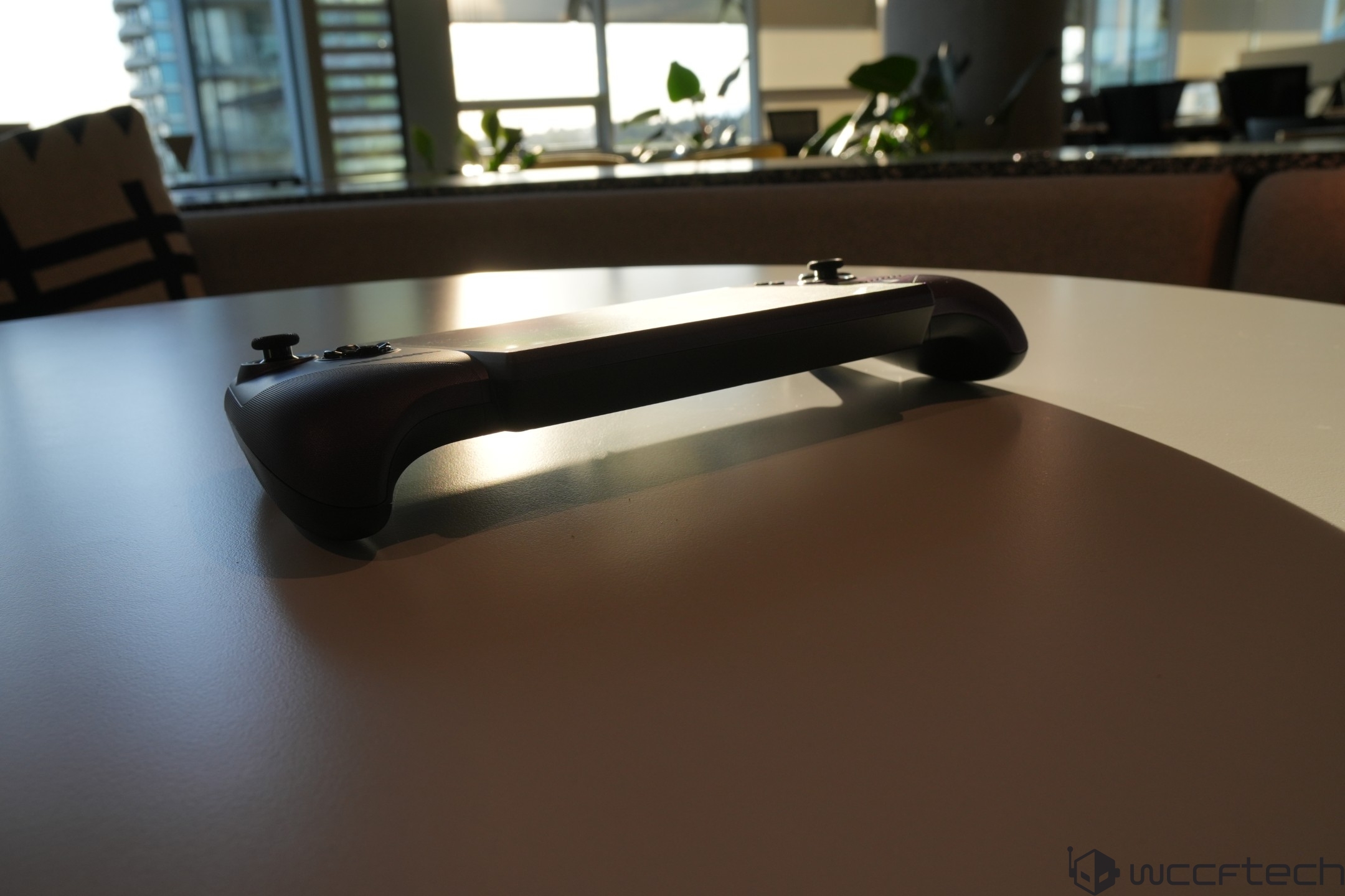 A handheld gaming console rests on a table in a sunlit room, showing side-mounted control grips and buttons.