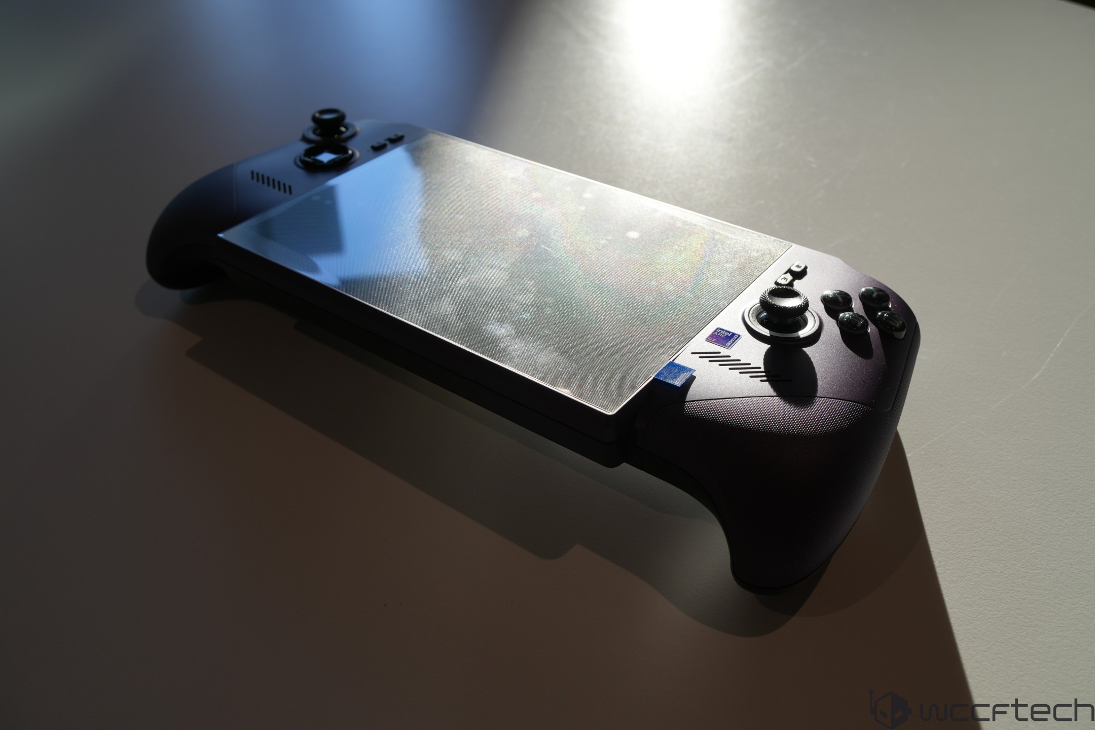 A handheld gaming device featuring dual analog sticks and several control buttons is placed on a table, with a distinct design emphasizing ergonomic grips and a tablet-like screen.