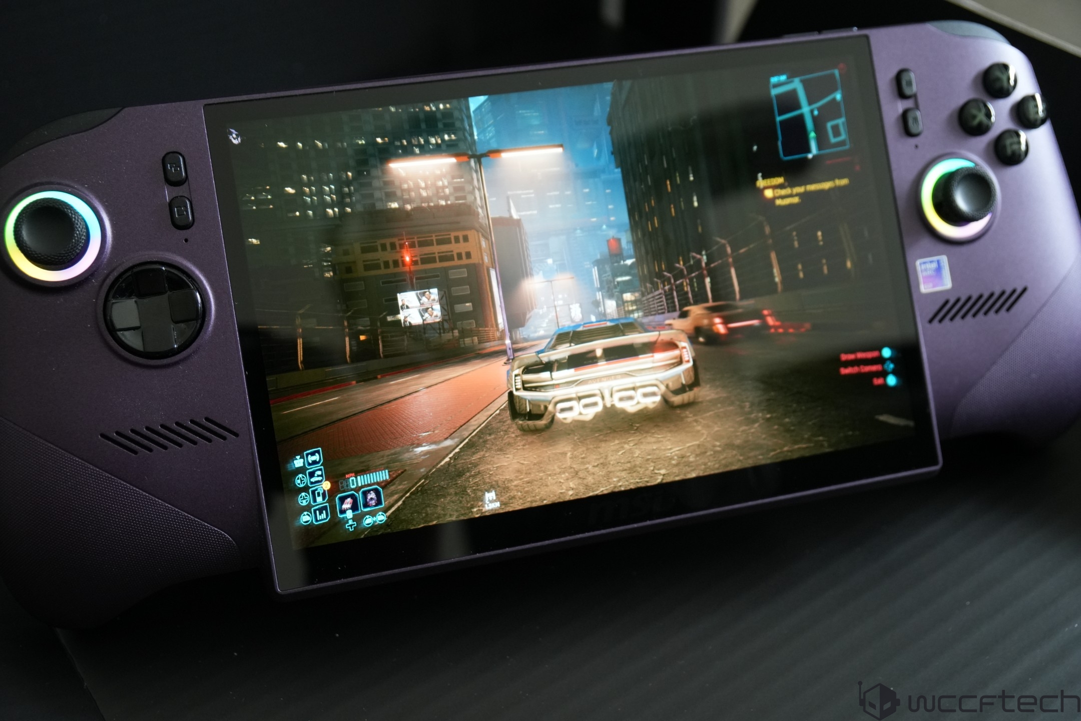 A gaming handheld displays a scene from Cyberpunk 2077 showing a car driving through a nighttime city street, with visible UI elements and a visible Intel sticker on the device.