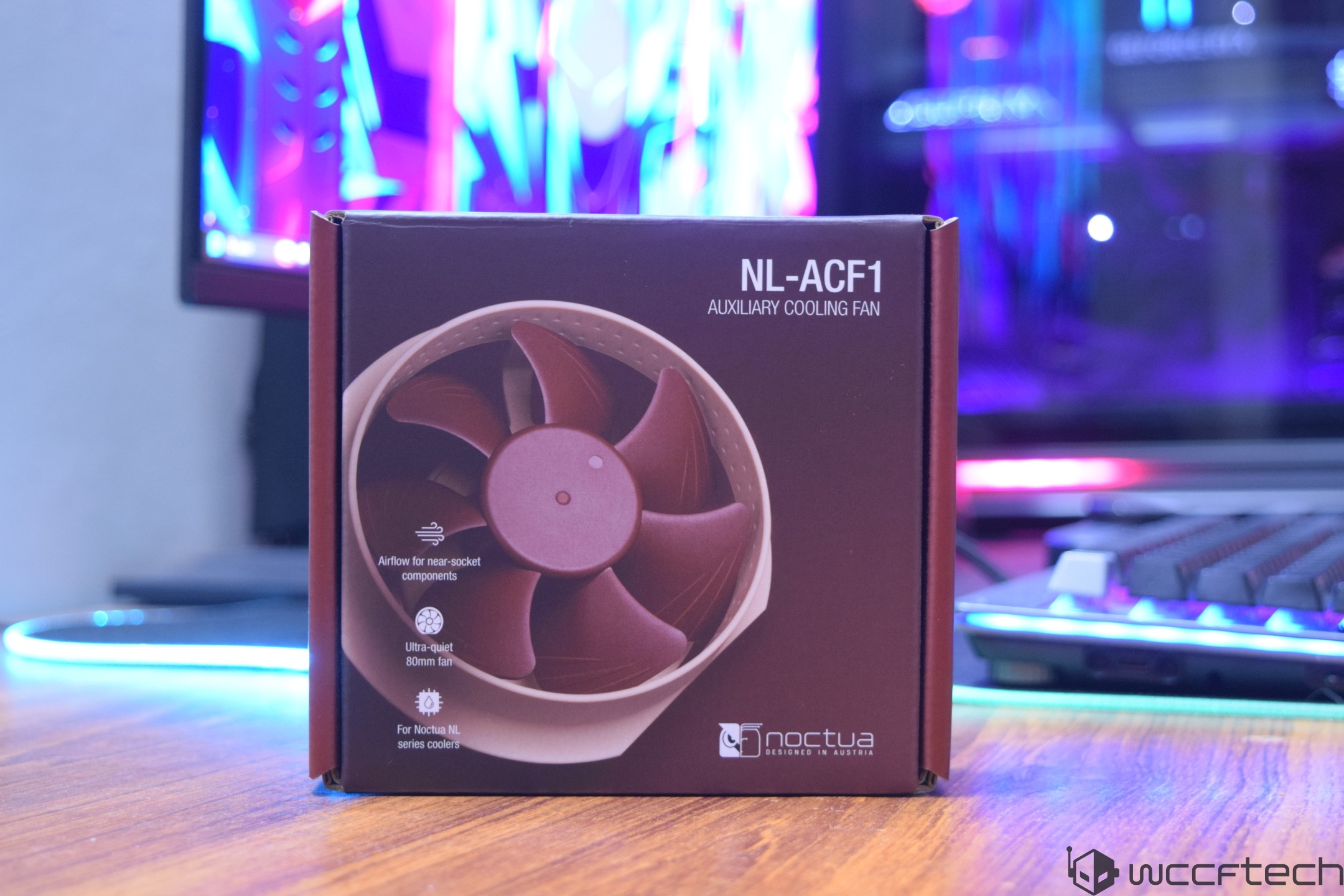 A Noctua NL-ACF1 auxiliary cooling fan box sits on a desk, featuring product highlights such as Airflow for near-socket components and Ultra-quiet 80mm fan.