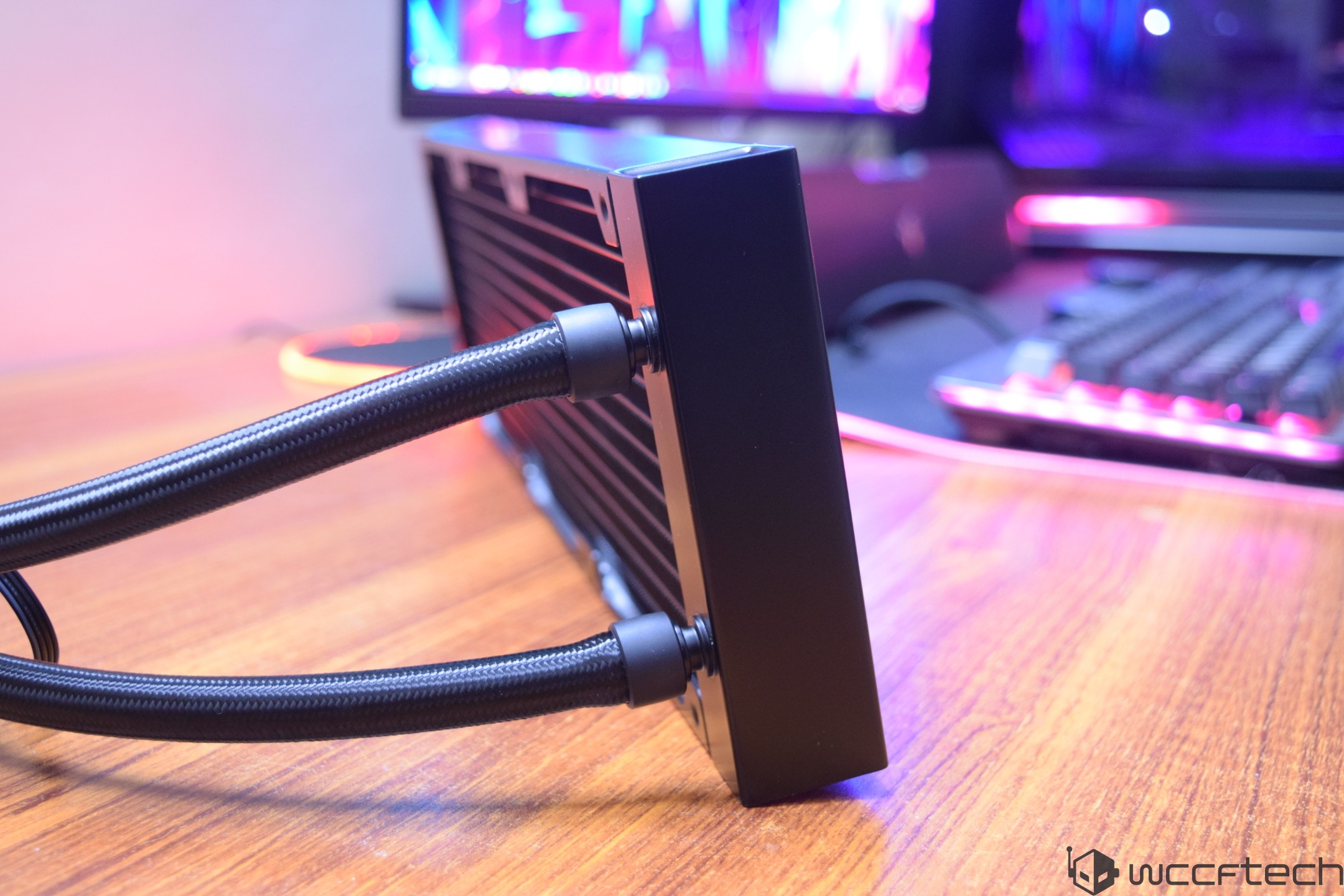 A close-up view of a black PC liquid cooling radiator and its braided hoses on a wooden desk, with RGB-lit components and a keyboard in the blurred background.