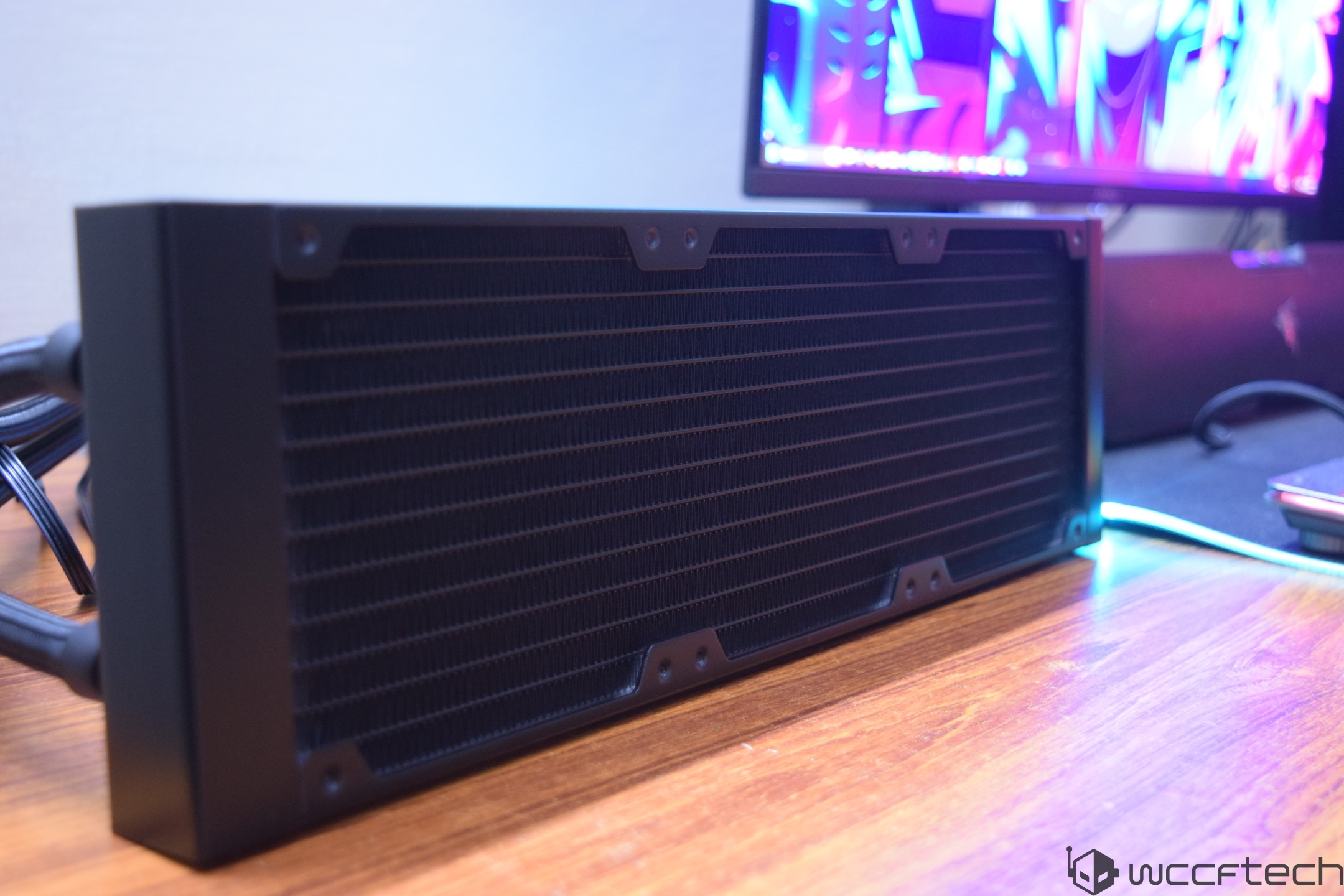 A desktop liquid cooling radiator is displayed on a wooden desk, with a monitor showing colorful abstract graphics in the background.