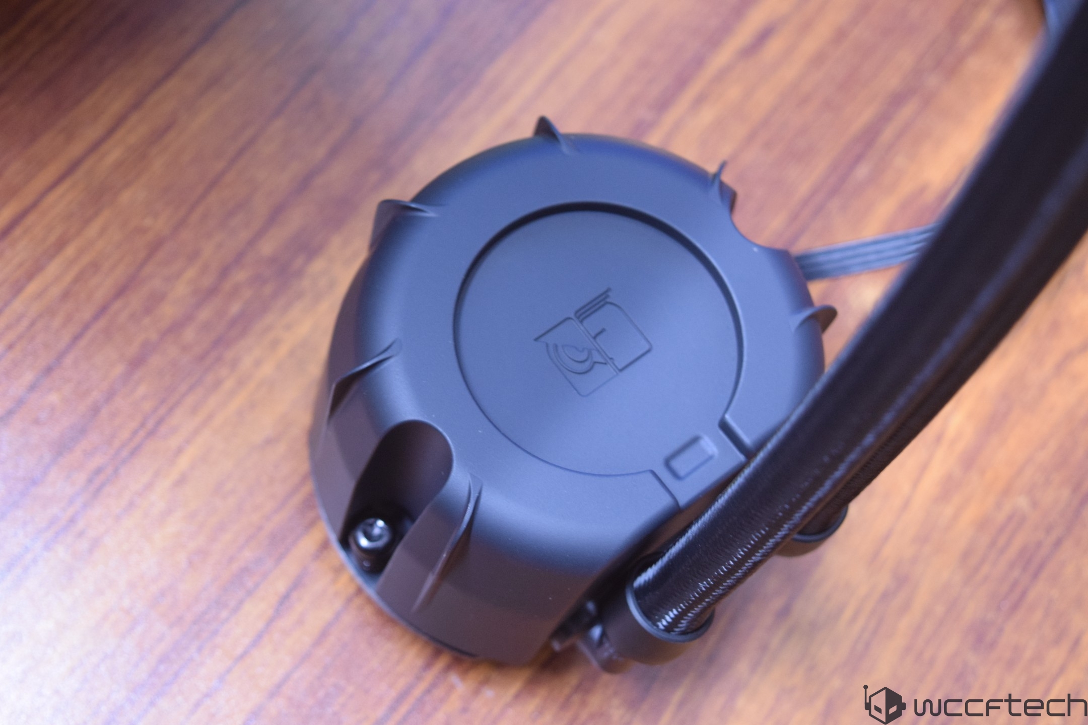 A close-up of a black AIO liquid cooler pump with stylized fan and wave icons on a wooden surface.