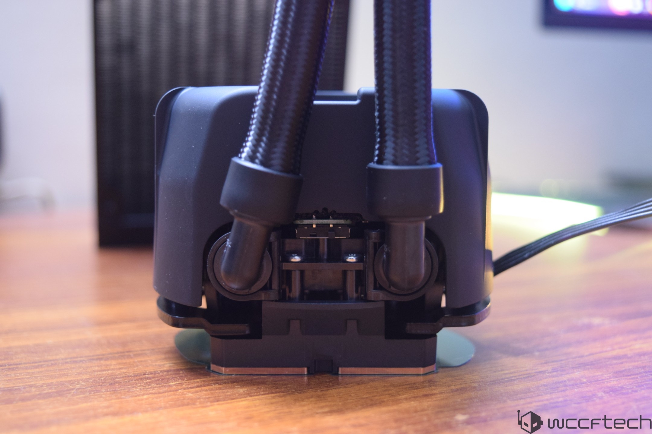 A close-up of a PC liquid cooling system's tubing and pump mounted on a wooden desk, with a blurred radiator in the background.