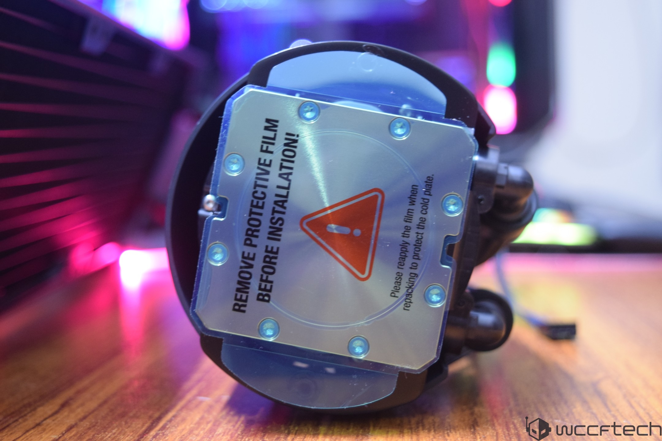 A liquid cooler with a clear warning label reading 'REMOVE PROTECTIVE FILM BEFORE INSTALLATION!' is displayed on a wooden surface.