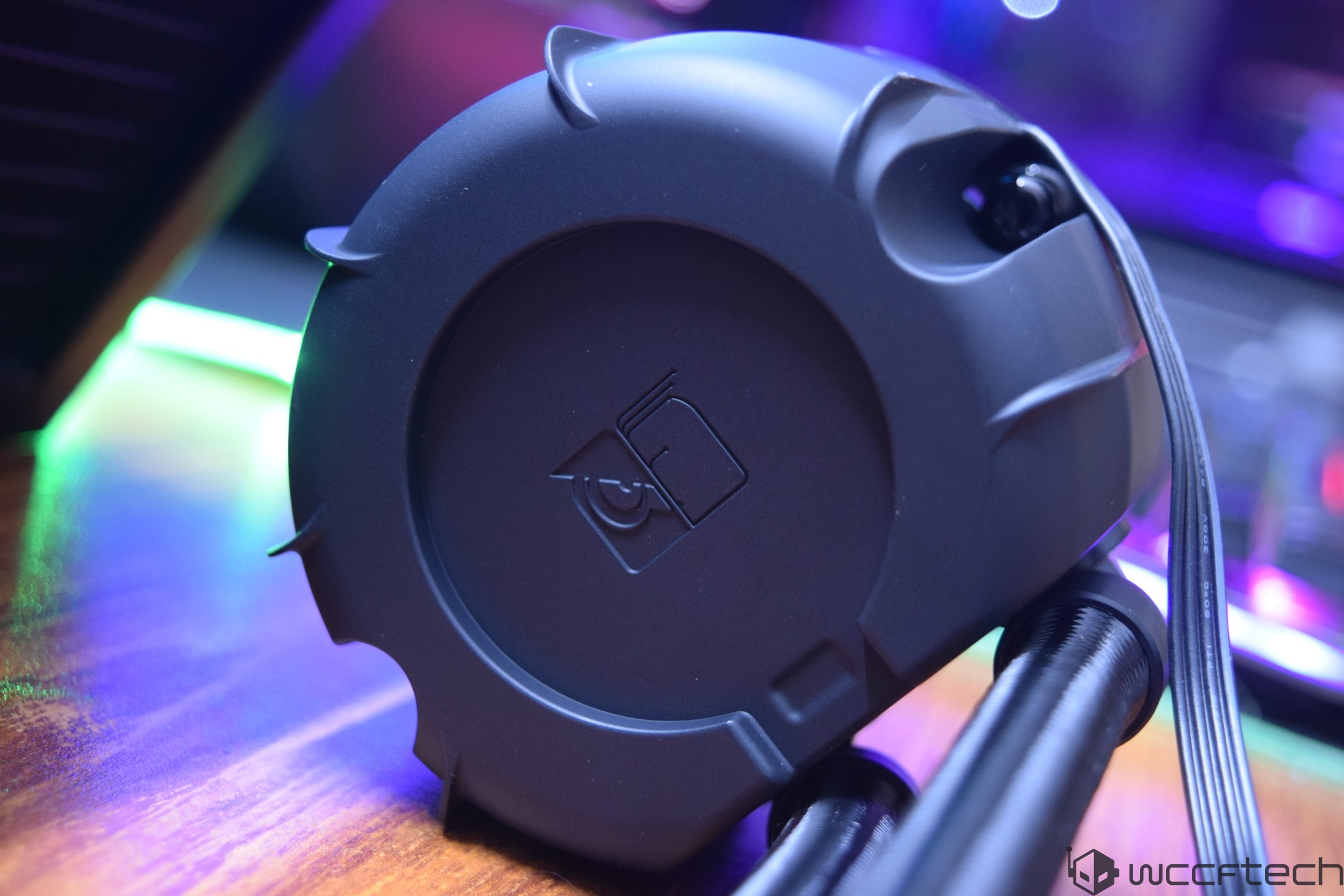 Close-up of an unbranded black liquid cooling pump with integrated cables, set on a desk with ambient purple and green lighting.