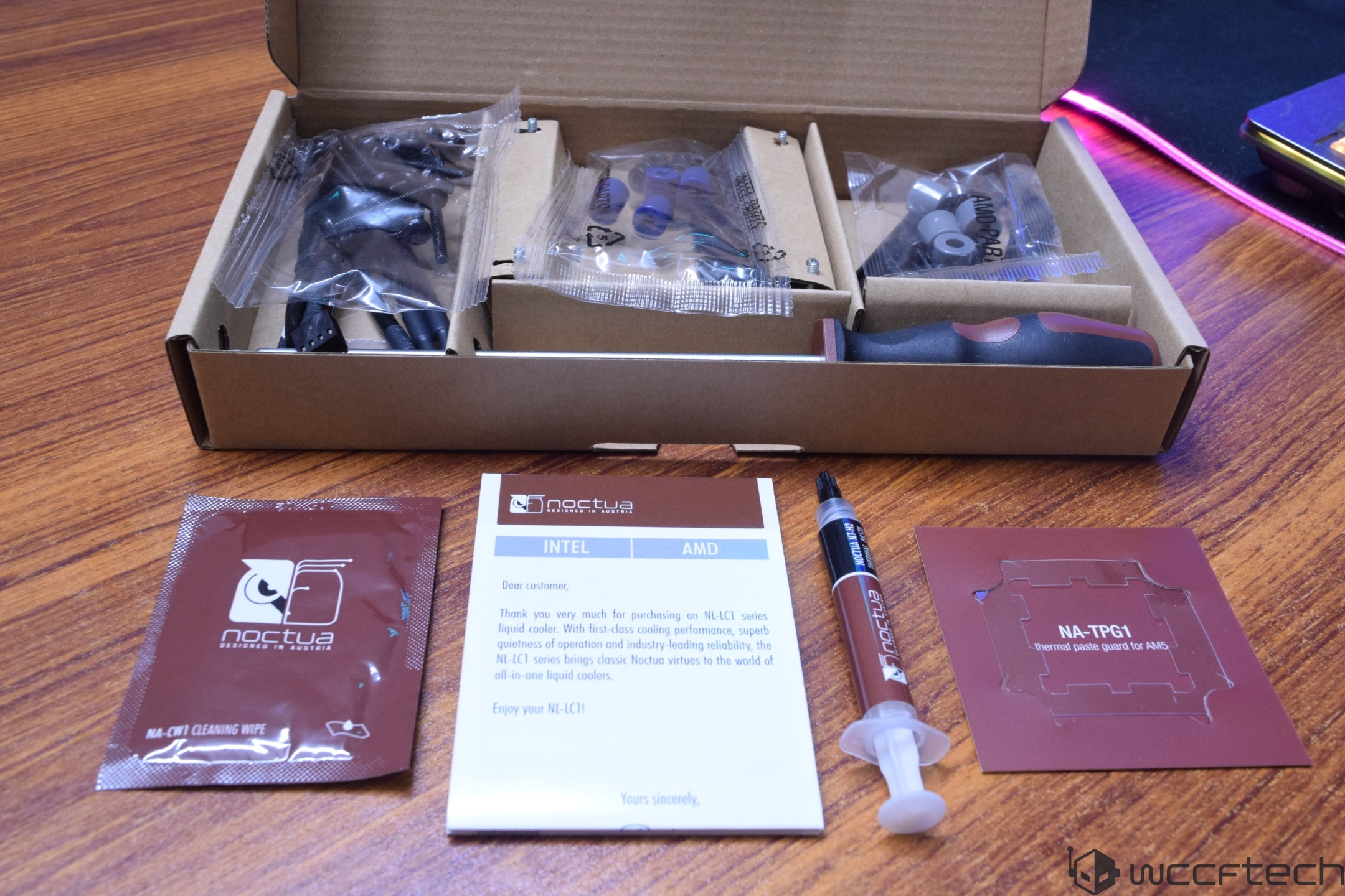 A Noctua NL-LC1 series liquid cooler installation kit is displayed with a screwdriver, thermal paste syringe, cleaning wipe, and instruction manual on a wooden table.