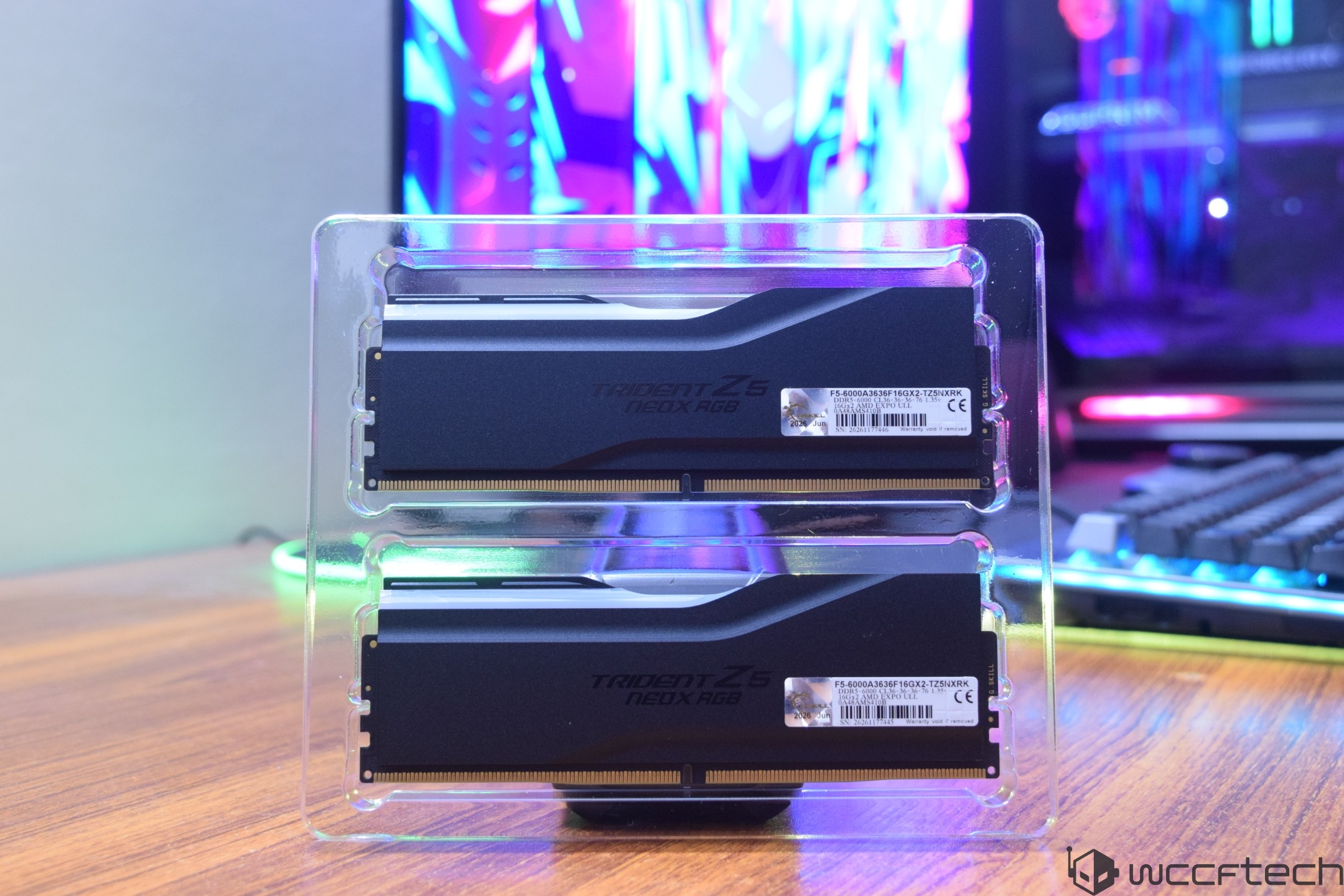Two G.SKILL Trident Z5 Neo X RGB RAM modules are displayed in transparent packaging on a desk with a colorful computer setup in the background.
