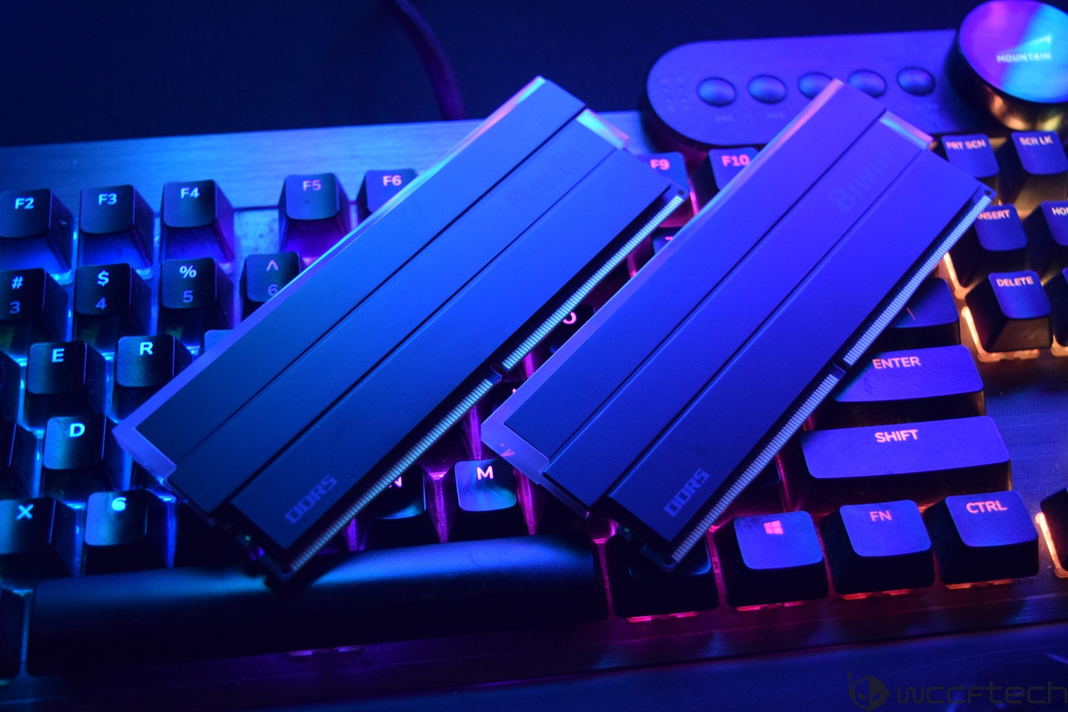 Two DDR5 RAM sticks lying on a backlit mechanical keyboard with visible QWERTY keys and a mountain logo in a dark blue and purple lighting environment.