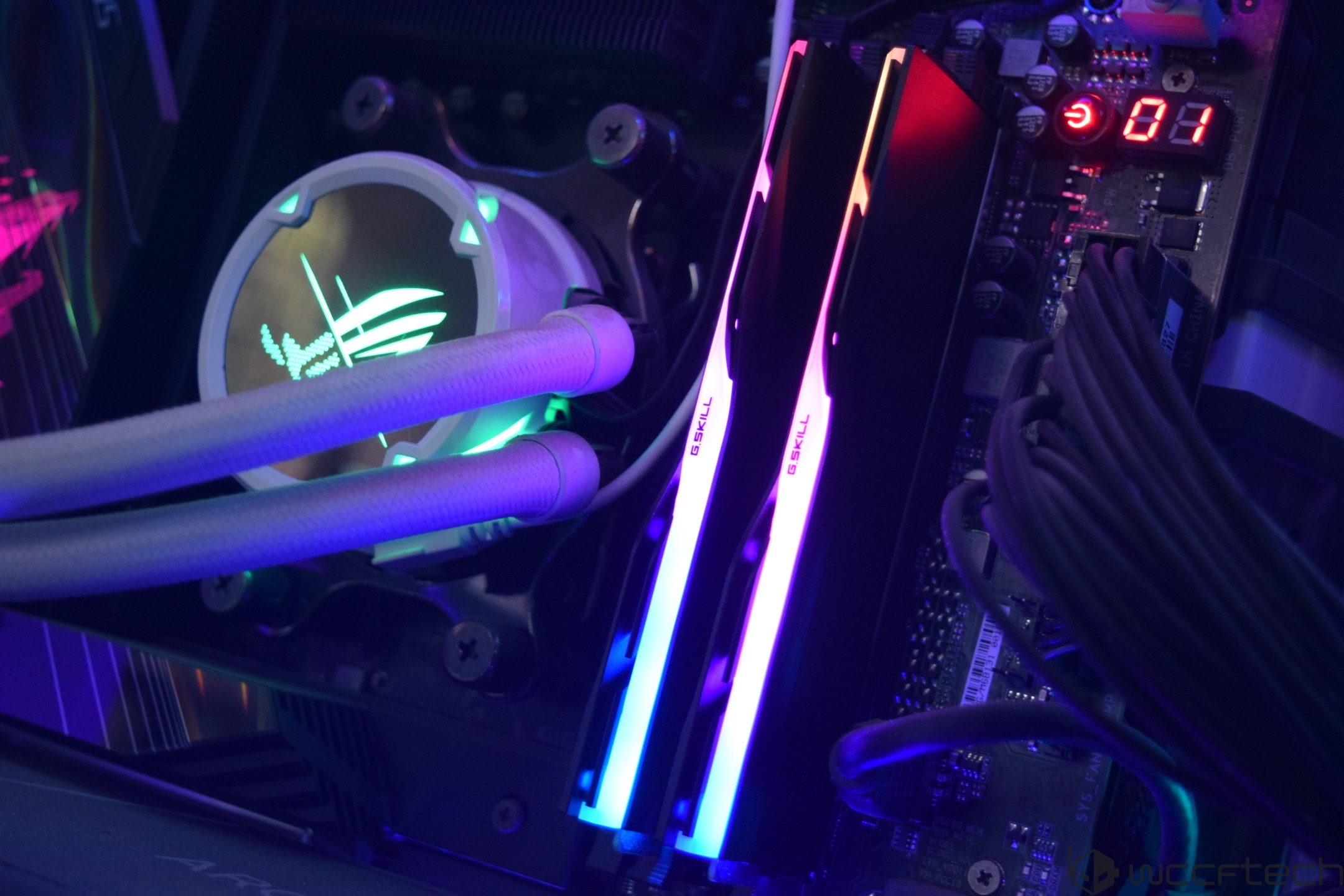 A close-up of a computer system shows two G.Skill RGB RAM modules and an illuminated ASUS cooler with the ROG Eye logo, alongside an 01 error code on the motherboard display.