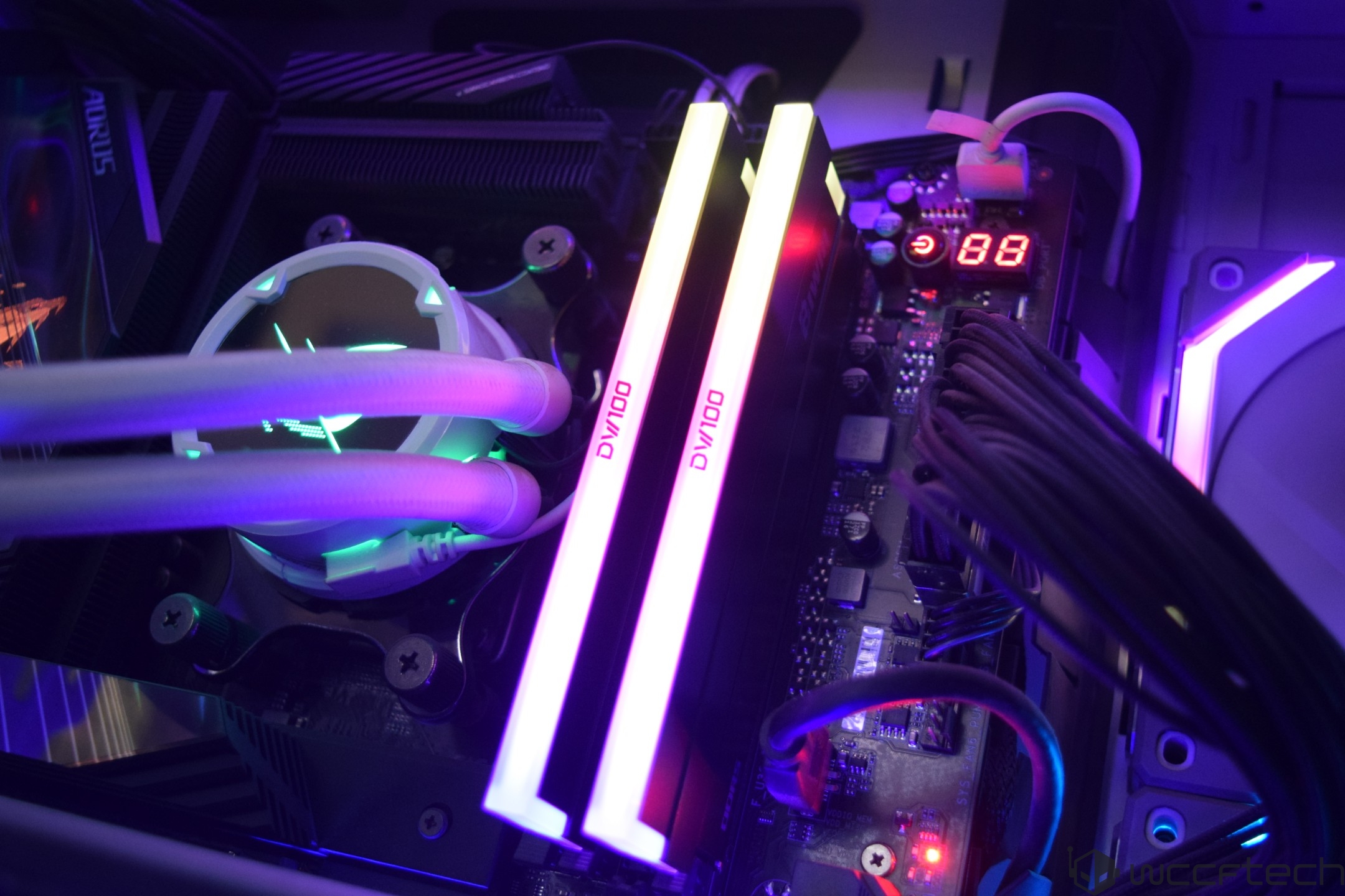 An illuminated internal view of a PC showcasing AORUS RAM sticks labeled DVI00 and a digital display reading '88,' alongside an RGB-lit liquid cooling system.