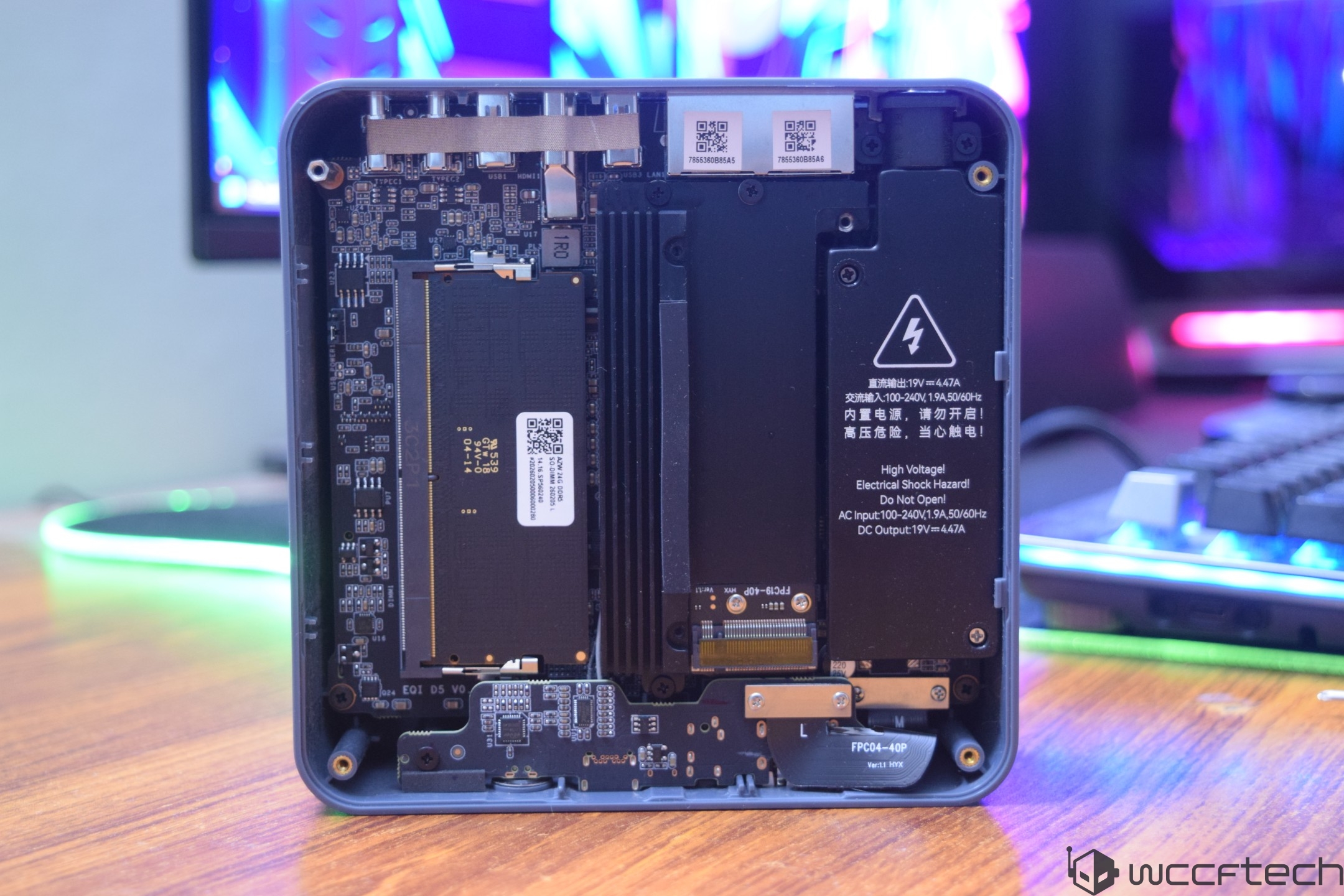 An open unbranded mini PC showing exposed internal components, including a RAM module and a warning label with 'High Voltage' text, placed on a wooden desk with a blurred background.