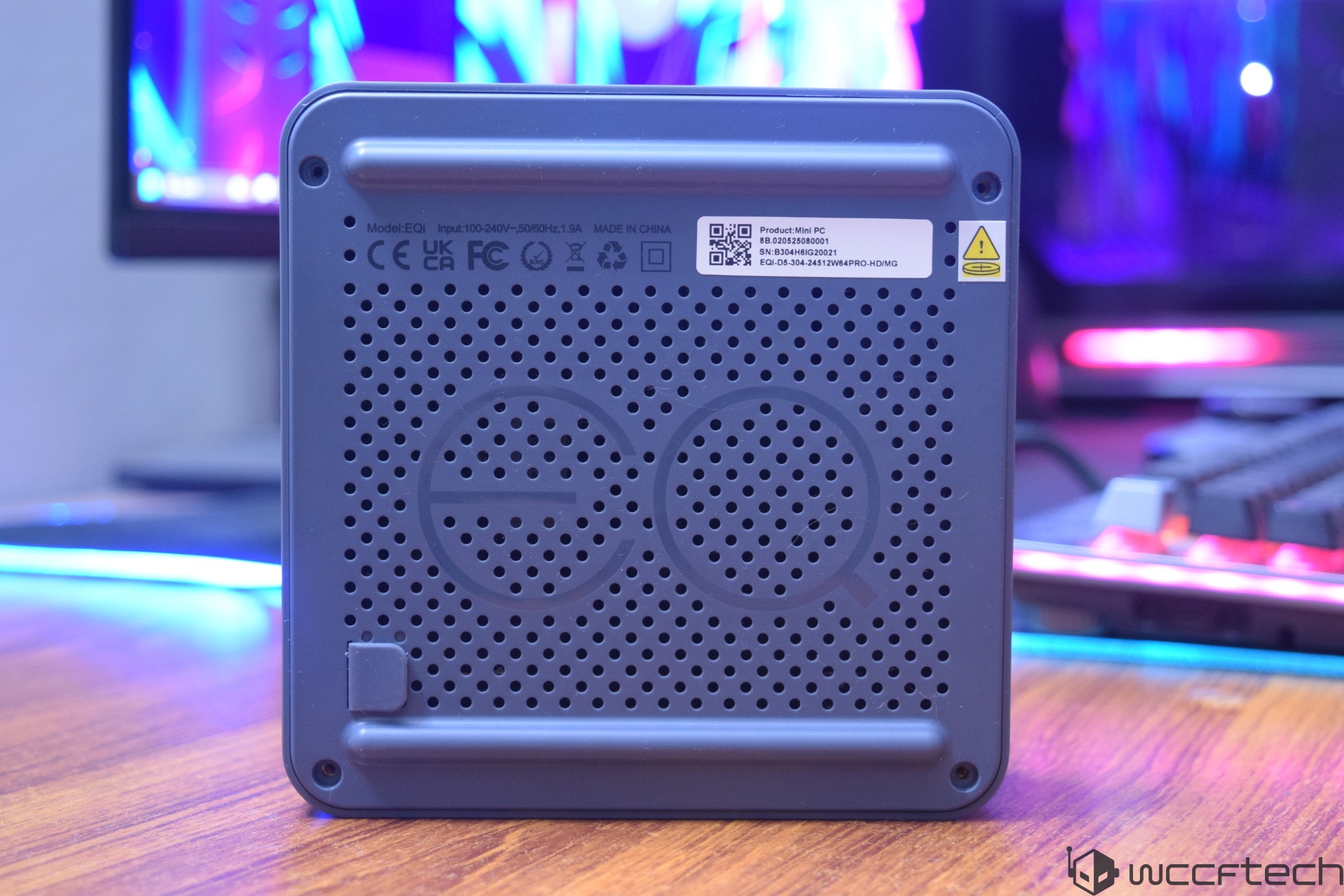 The back of a mini PC labeled Model: EQI with various certifications, a QR code, and a warning symbol, placed on a desk with blurred computer screens in the background.