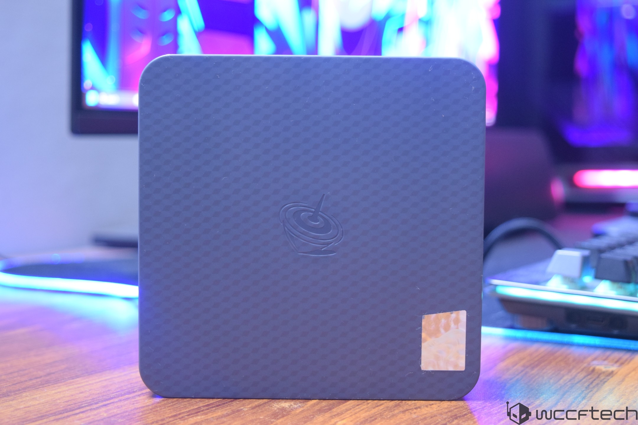 A small, dark-colored device with a textured pattern on the top, featuring an embossed logo resembling a stylized spinning top, placed on a wooden surface with a blurred monitor displaying colorful graphics in the background.