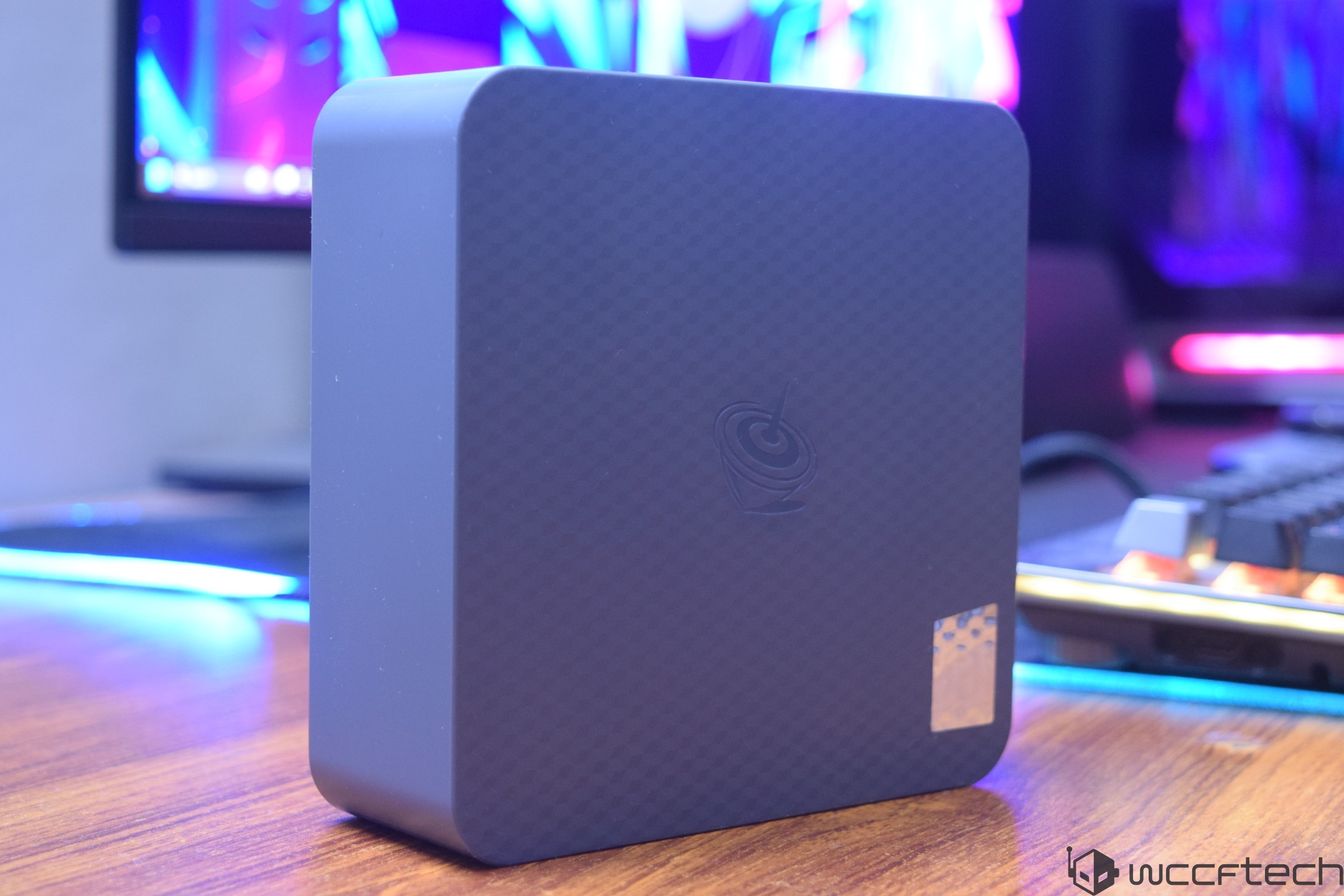 A compact, dark blue mini-PC with a subtle hexagonal pattern and an embossed logo, placed on a wooden desk with a colorful LED-lit keyboard and monitor in the background.