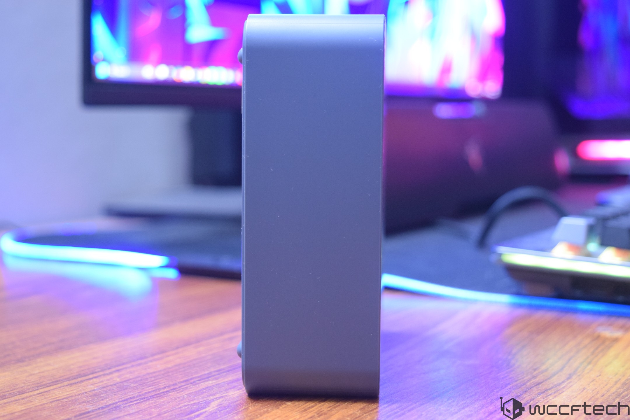 A vertical unbranded desktop PC case is positioned on a wooden desk with a computer monitor and keyboard in the background, illuminated by RGB lighting.