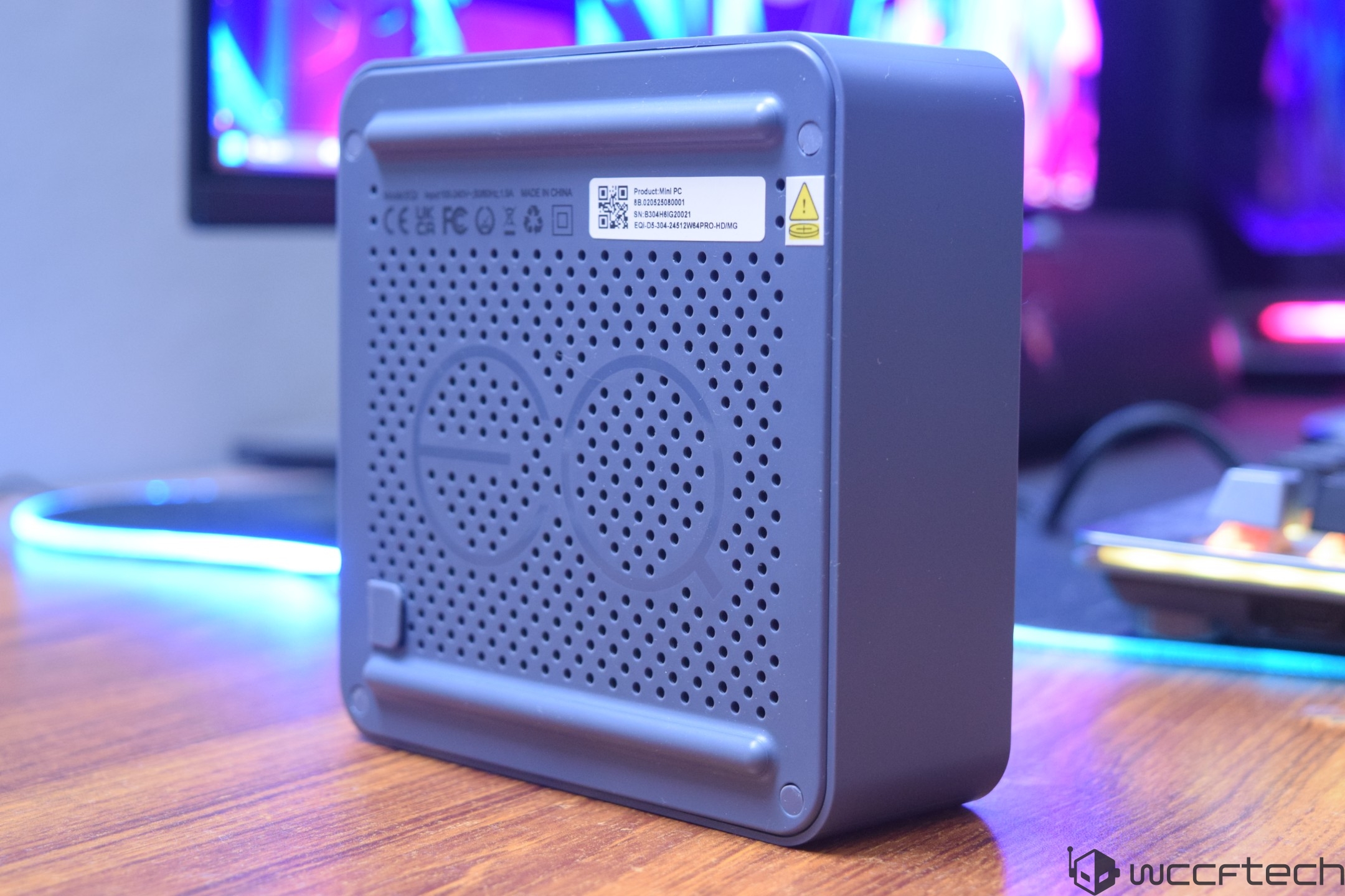 A gray EQ-branded mini PC is positioned on a wooden desk, with a monitor and keyboard visible in the background.