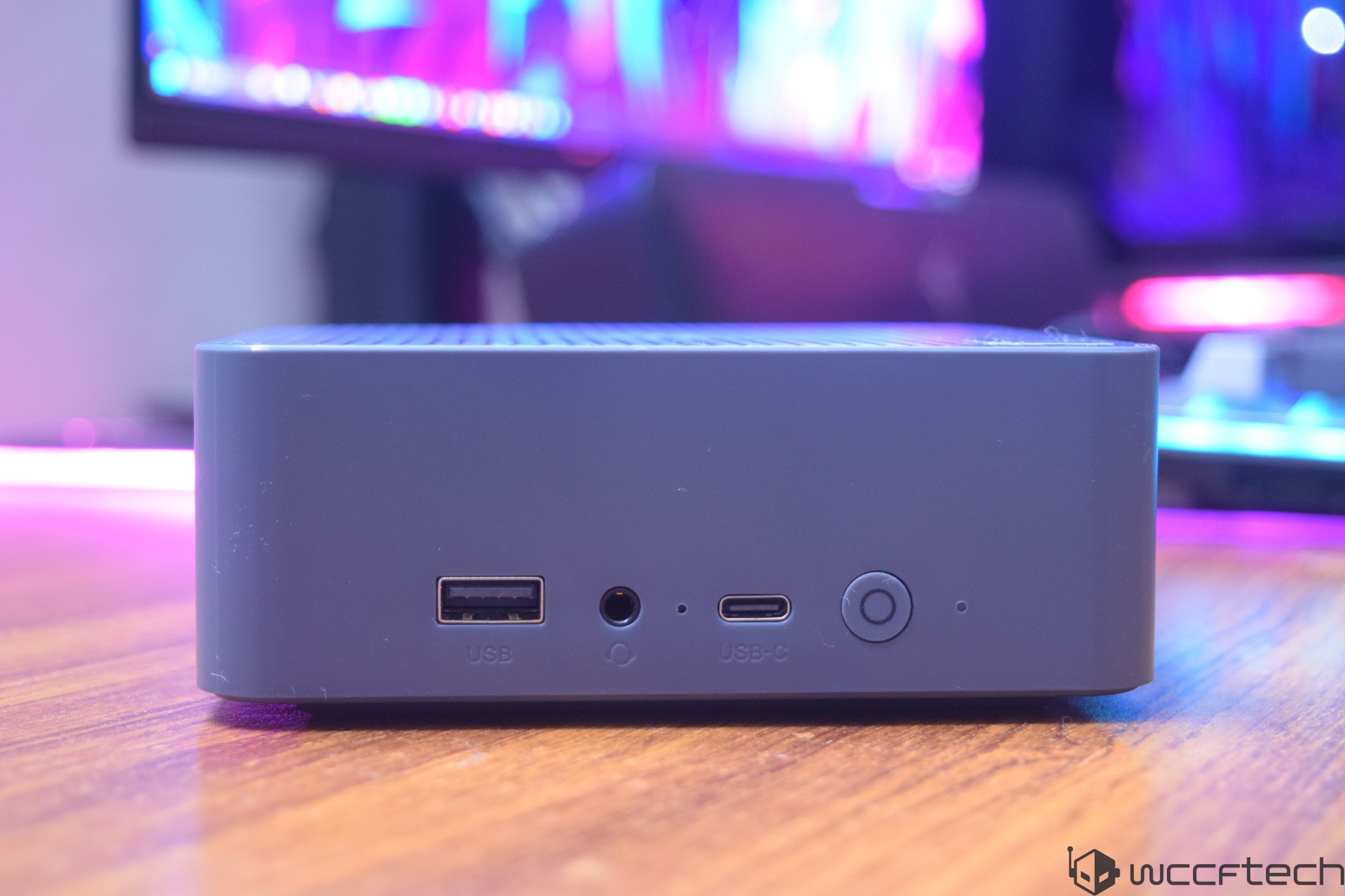A compact silver device with USB, USB-C, and headphone ports sits on a wooden surface in front of a blurred, colorful background screen.