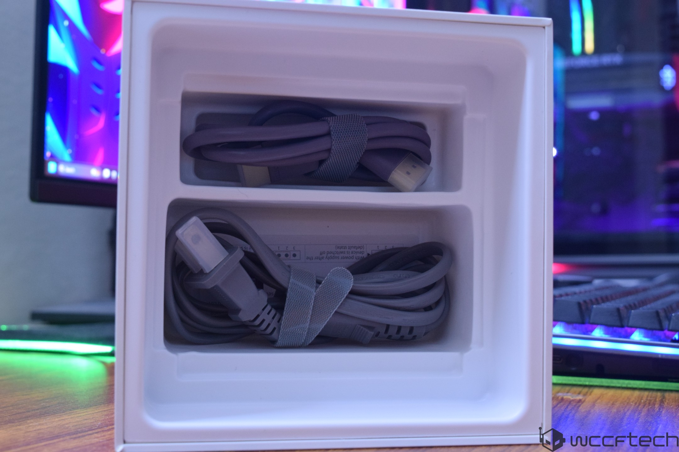 A white packaging box contains two coiled sets of unbranded cables with connectors, positioned in front of a computer monitor and a keyboard with colorful lighting.