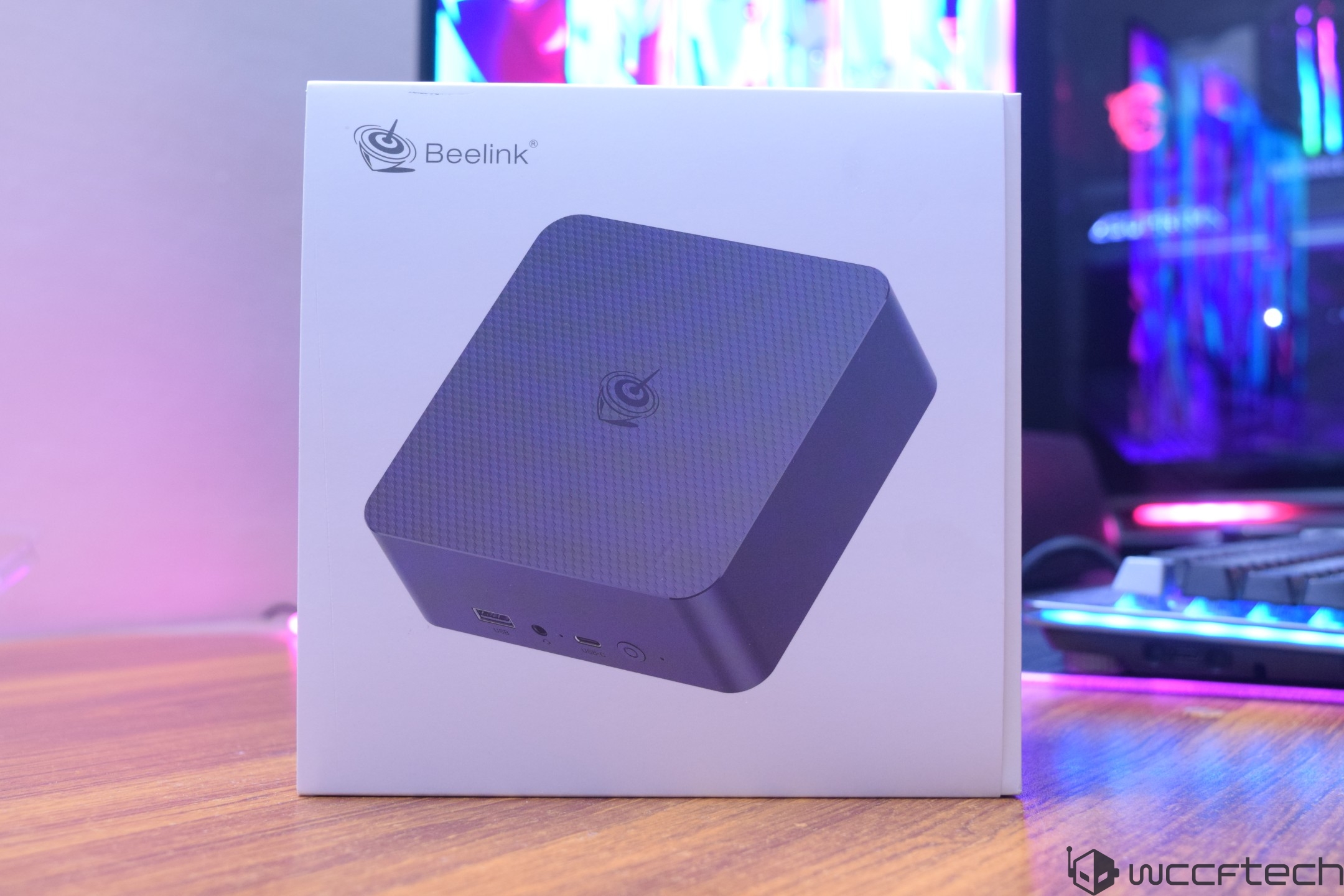 A Beelink mini PC box is displayed on a wooden table, with colorful LED lights glowing in the background.