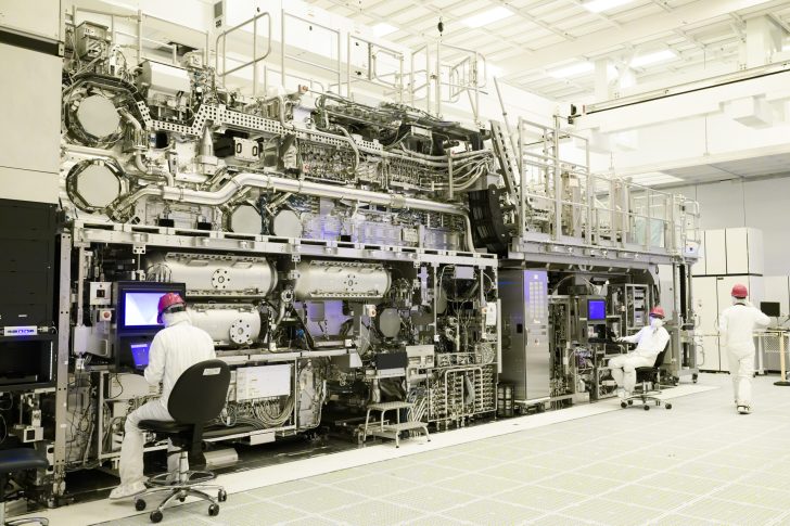 Technicians in a clean room work on an advanced ASML lithography machine with multiple screens and intricate machinery components.