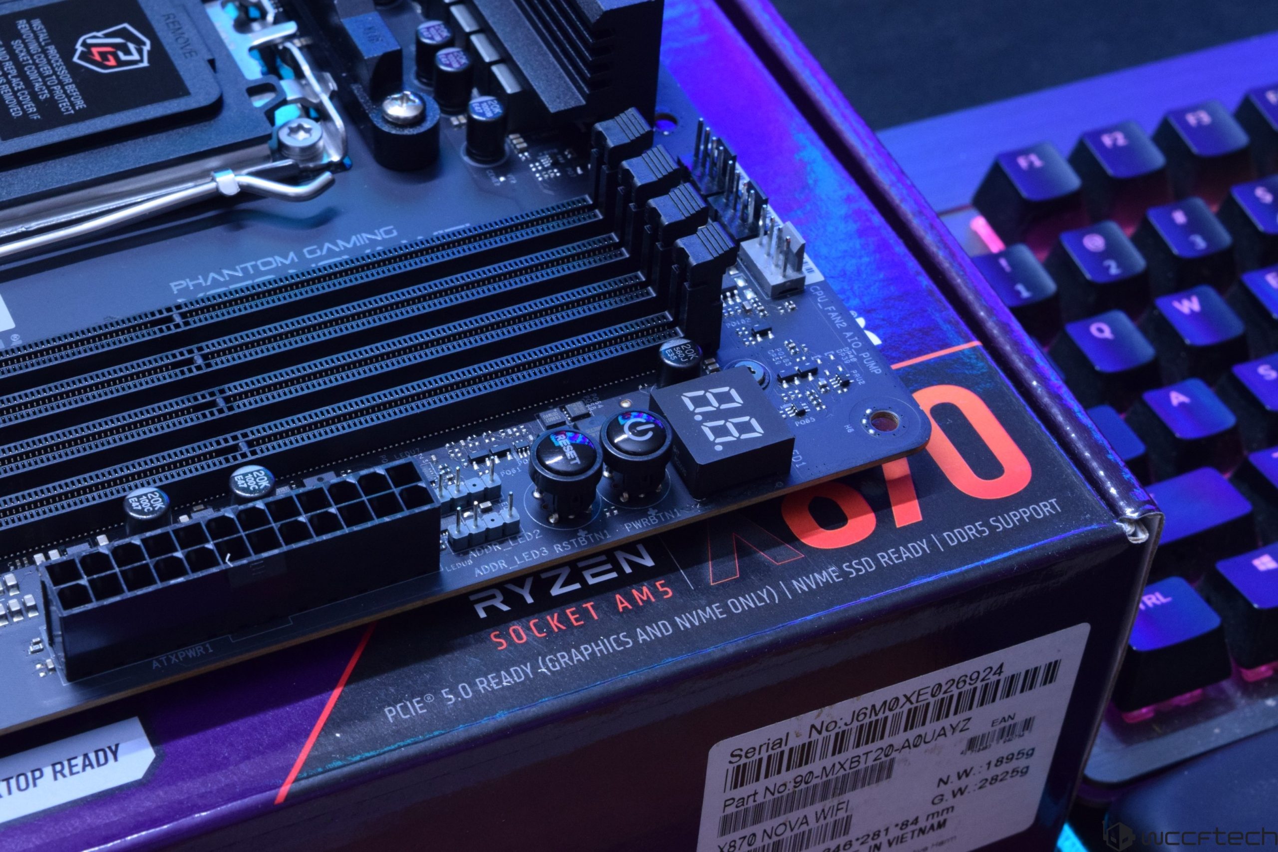 A close-up of an ASRock Phantom Gaming motherboard on a box labeled 'RYZEN SOCKET AM5' with visible power and reset buttons next to a digital display.