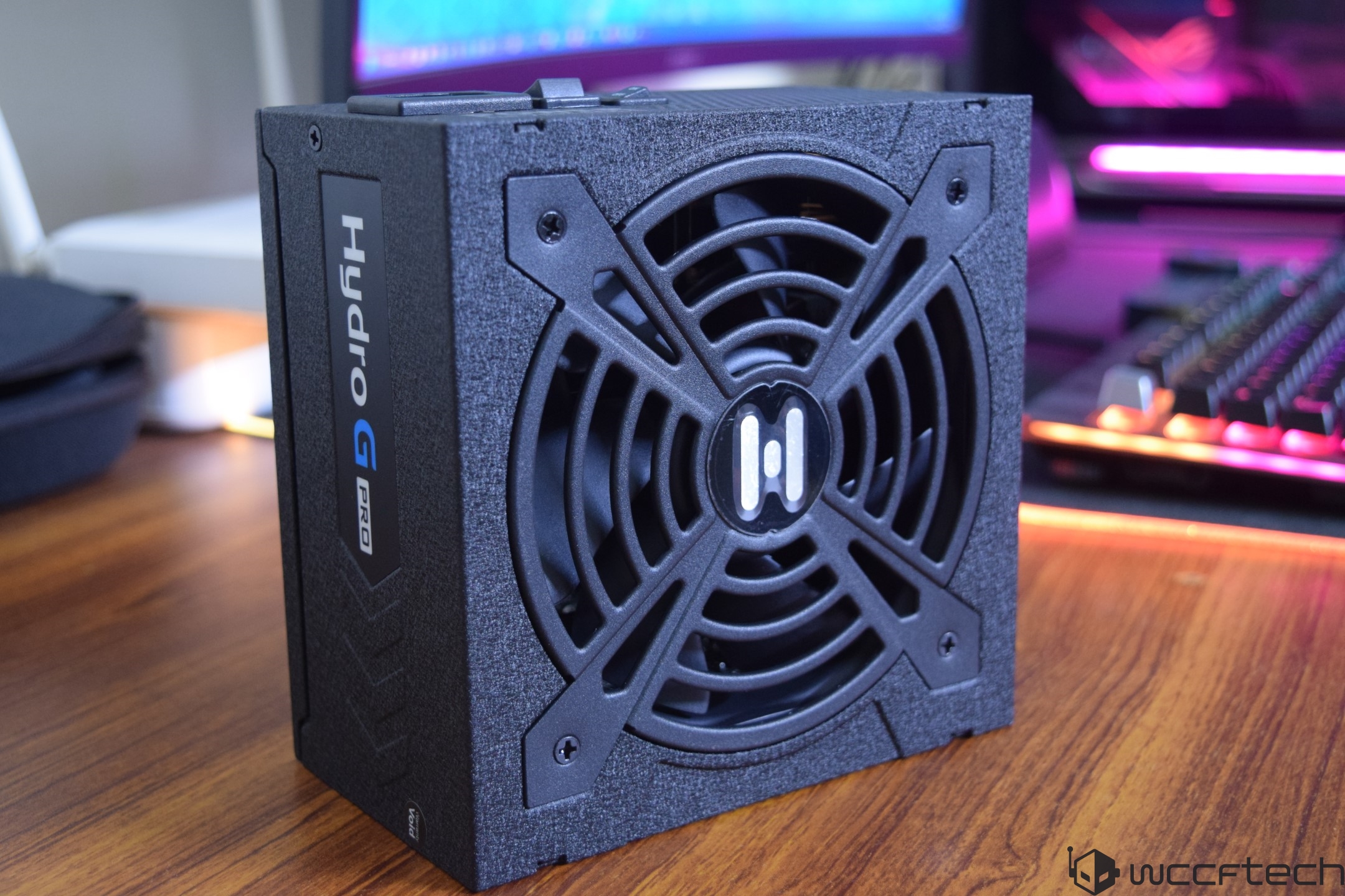 The image shows an FSP Hydro G Pro power supply unit on a wooden desk with a gaming setup in the background.