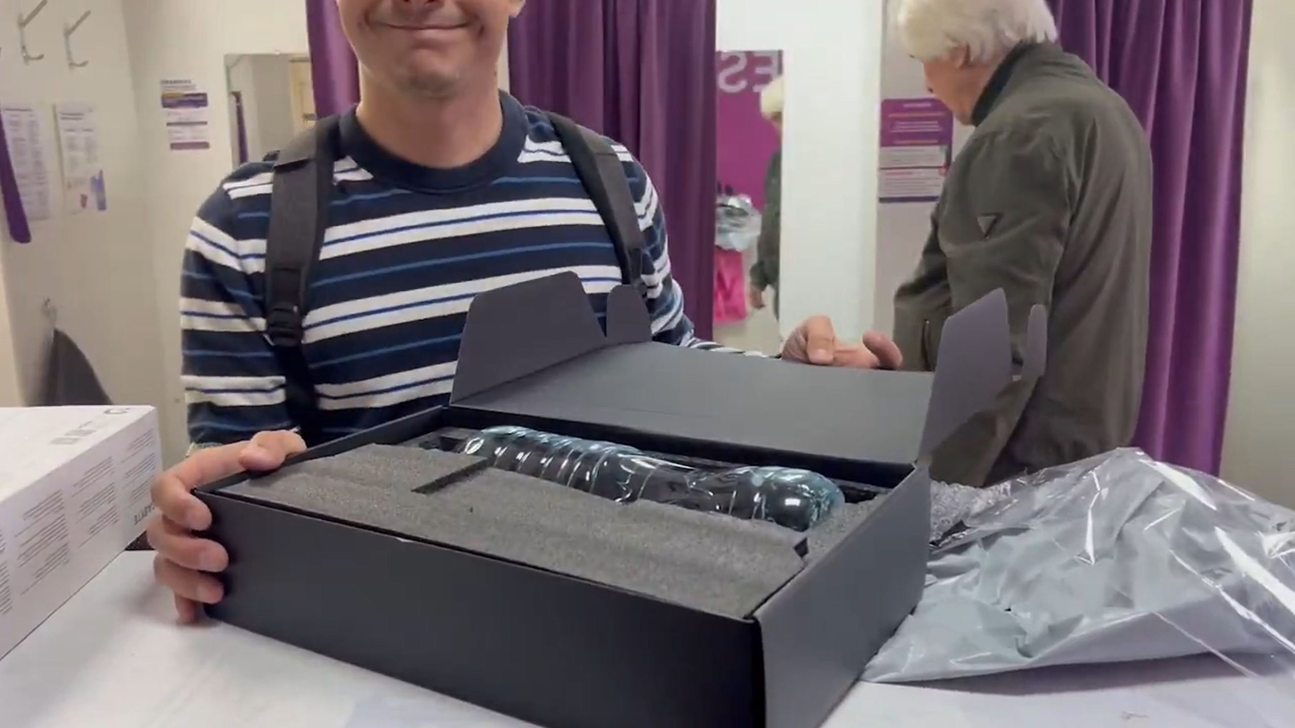 A person in a striped shirt unboxes a transparent bottle in a black foam-padded package, while another person stands in the background near purple curtains.