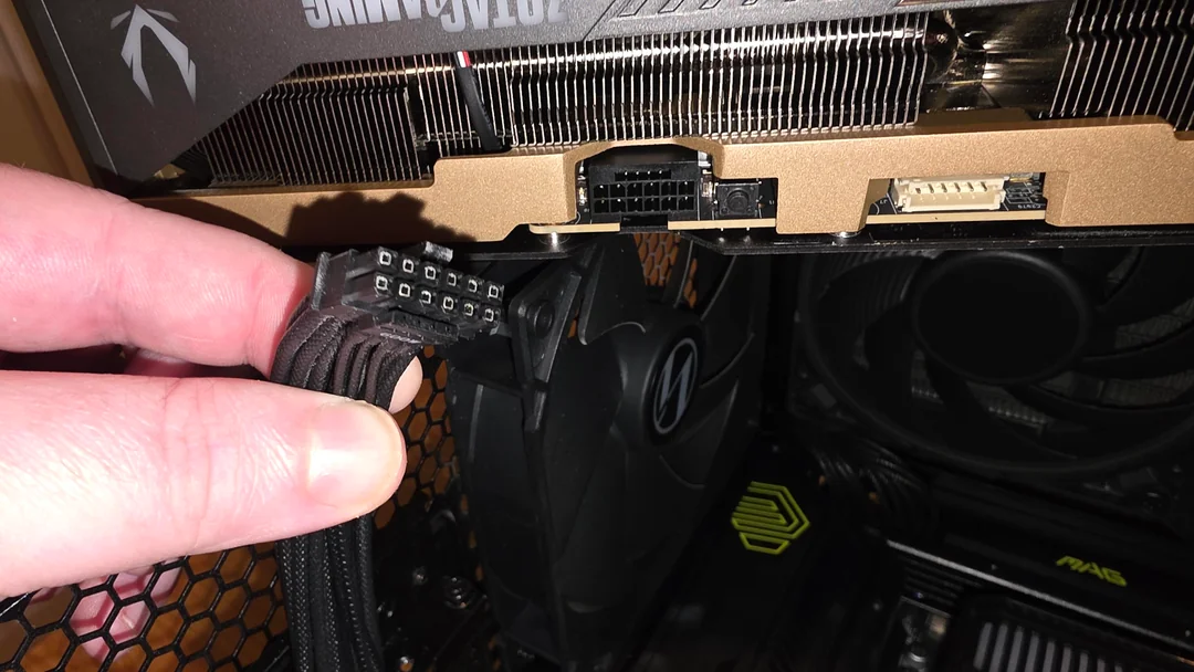 A hand holding an 8-pin power connector near the power port of an NVIDIA graphics card inside a PC case, with a visible cooling fan and branding on the card above.