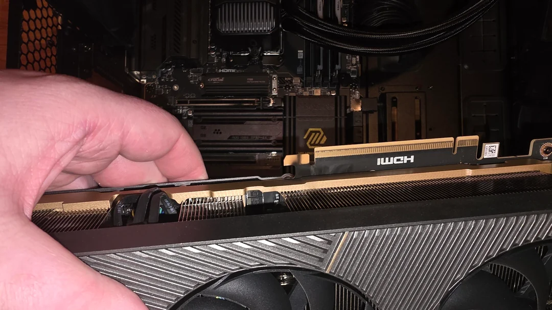 A hand is installing a NVIDIA GeForce graphics card into a motherboard inside a computer case, with visible RAM sticks labeled Crucial and a logo on the motherboard.
