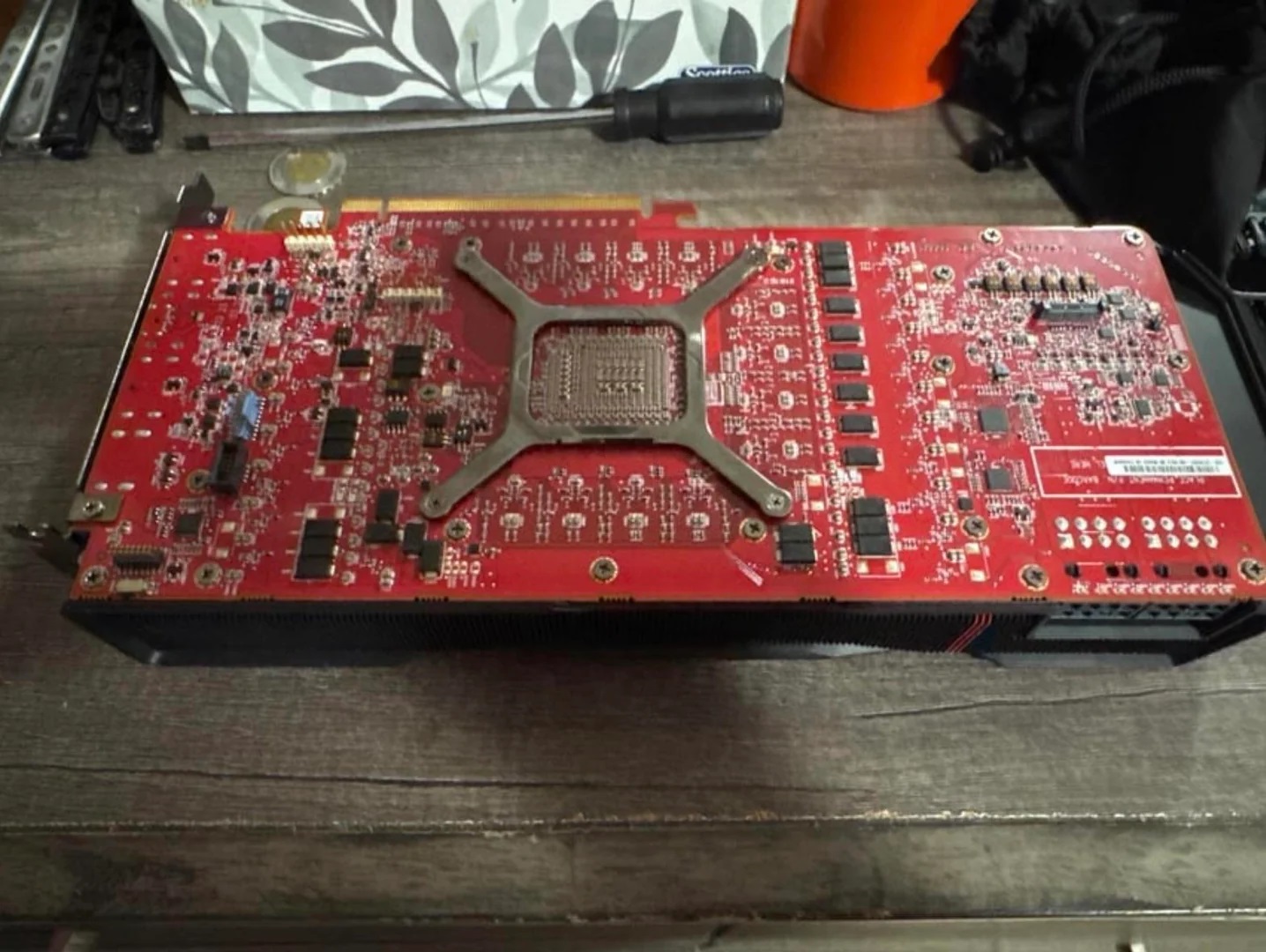 A red unbranded graphics card is placed on a wooden table with visible circuitry and a metal bracket in the center.
