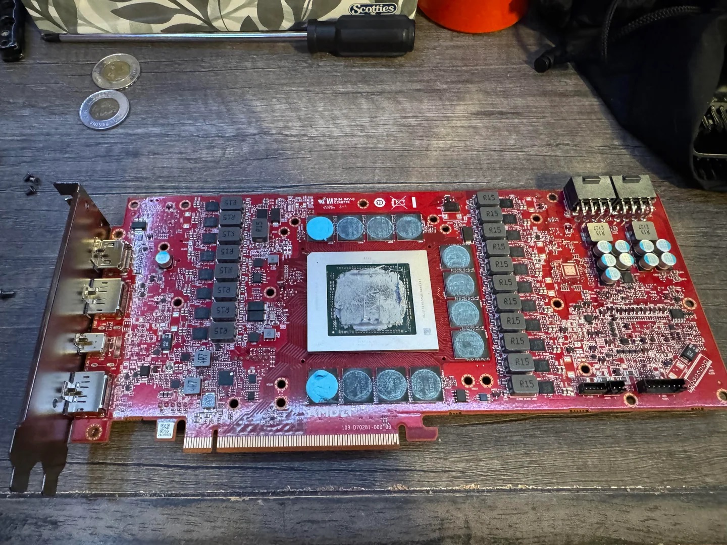 A red AMD graphics card with visible GPU chip and multiple capacitors lies on a wooden table next to a screwdriver and coins.