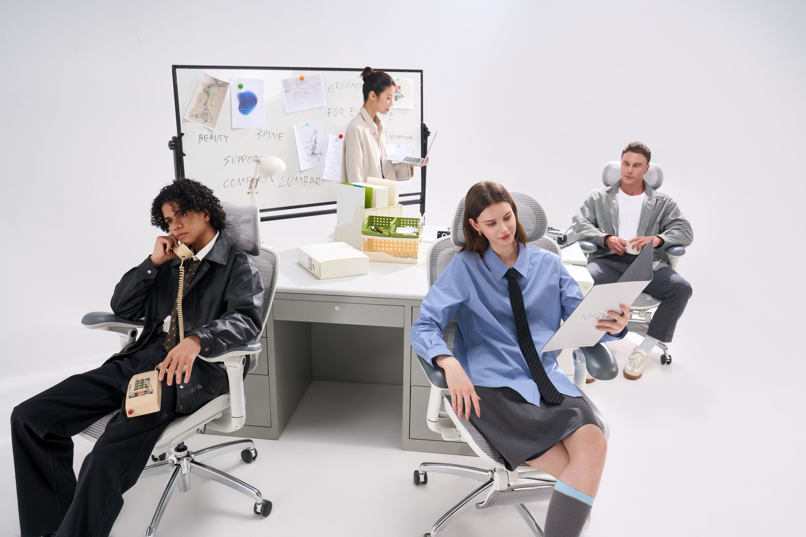 A group of people in an office setting, sitting on ergonomic chairs around a desk, with a whiteboard in the background displaying notes such as 'spine' and 'comfort lumbar'.
