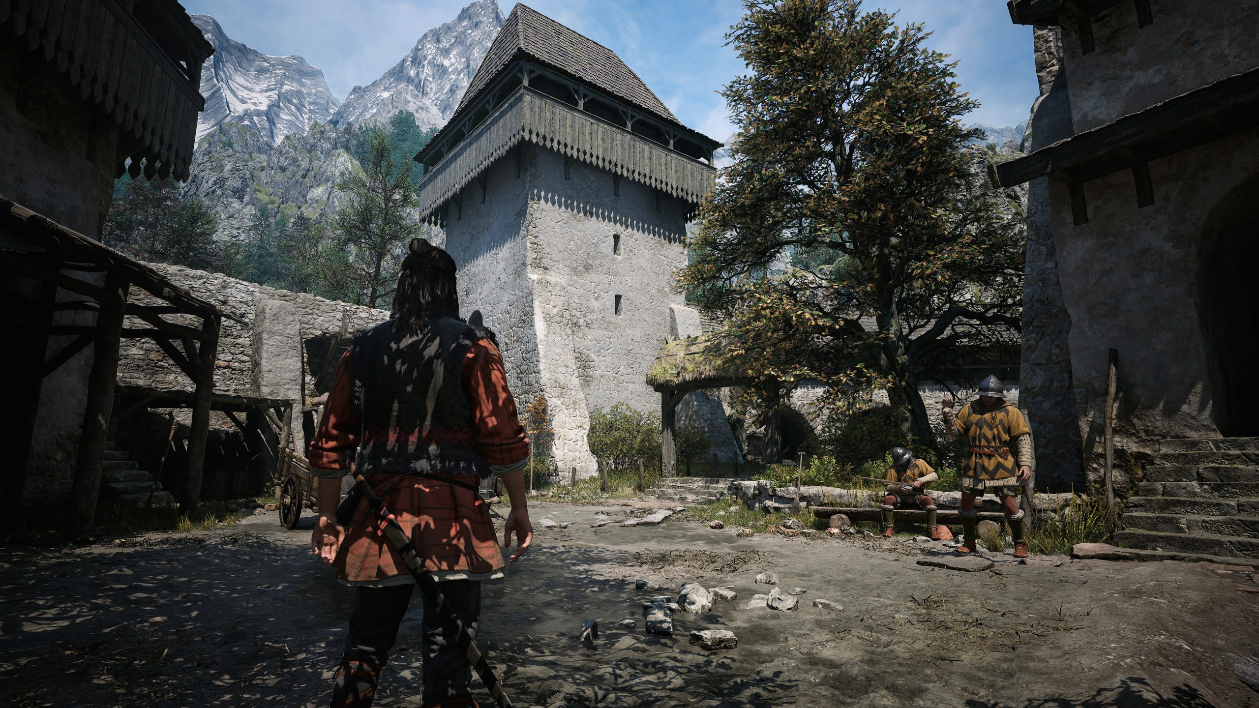 A character in medieval attire stands in a rustic courtyard with stone buildings, mountains in the background, and two armored figures sitting nearby in a scene from a video game.