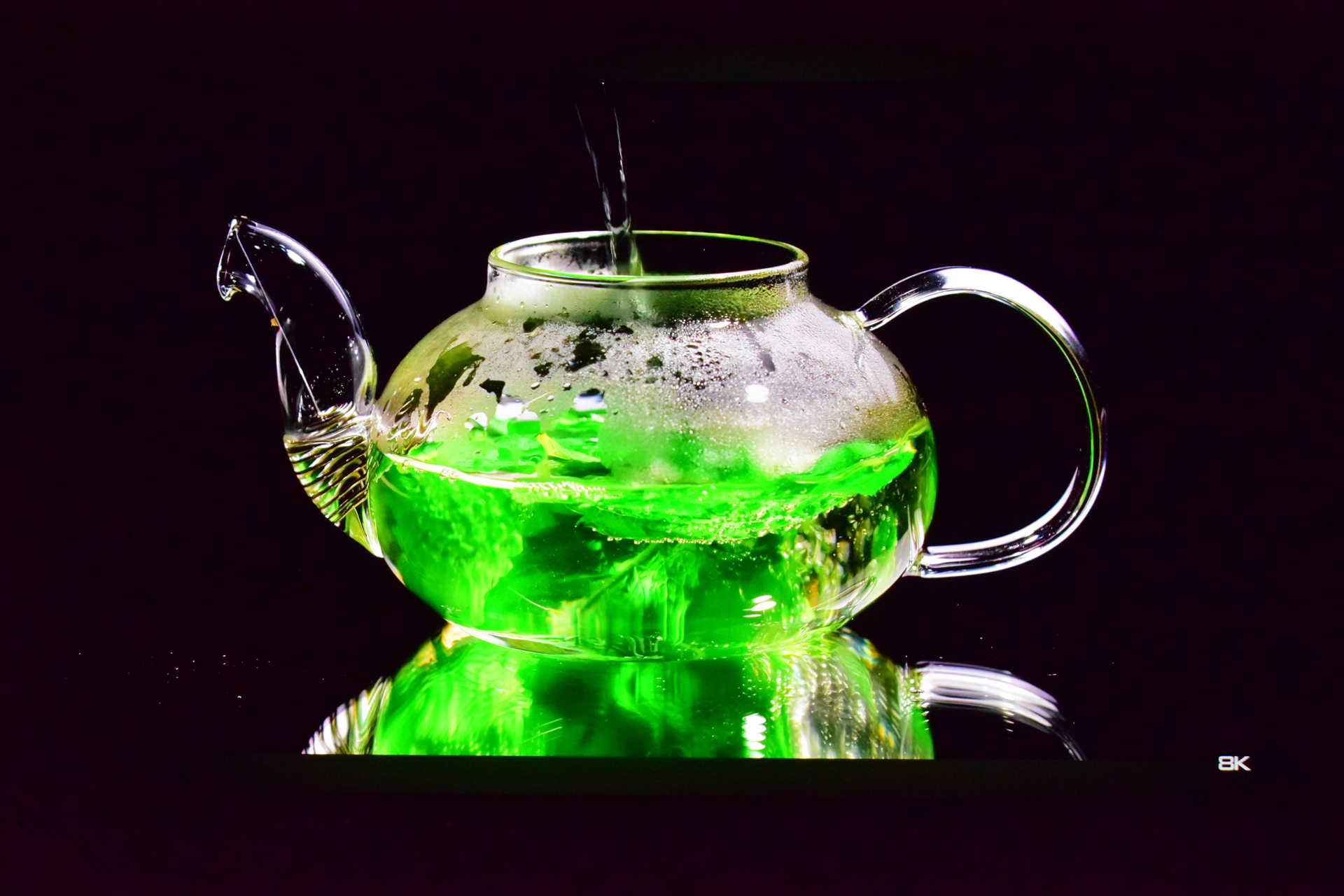 A clear glass teapot is being filled with steaming hot water, causing bright green liquid inside to swirl, with the text '8K' visible in the corner.