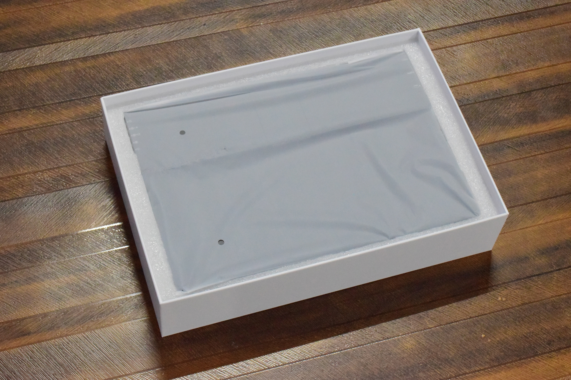 A cardboard box contains an unbranded electronic device wrapped in protective plastic packaging on a wooden floor.
