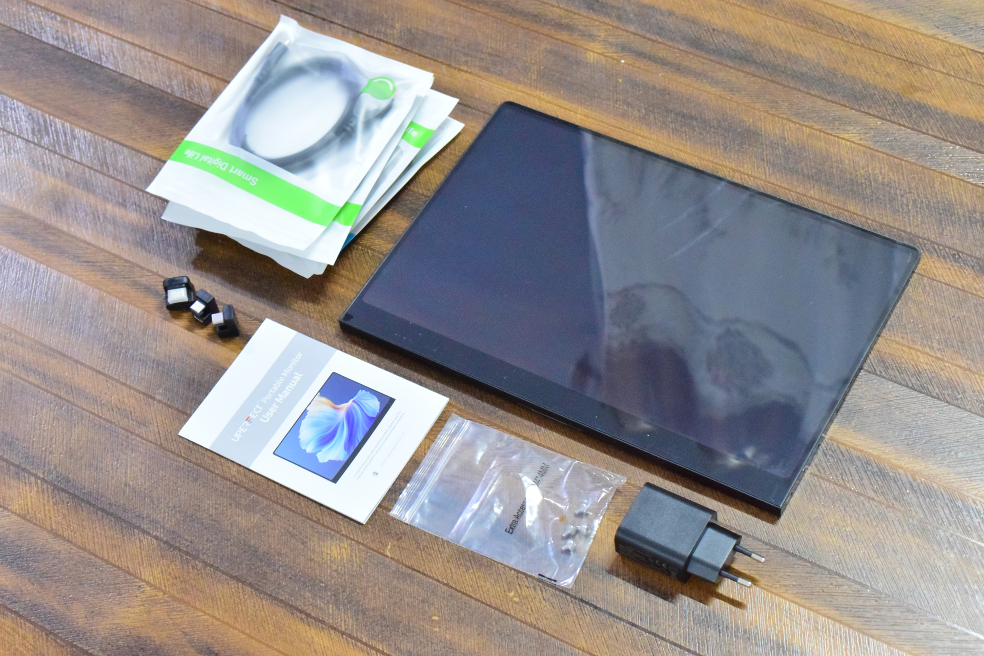 A UPerfect portable monitor and its accessories, including a charger, cables in labeled Smart Digital Life packaging, a user manual, and small plugs, are arranged on a wooden surface.