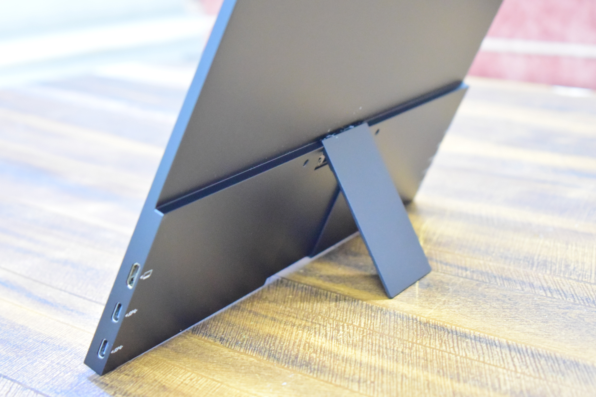 The back of an unbranded portable monitor on a wooden table, featuring a stand and three visible USB ports.