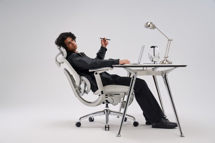 A person reclines in a white sihoo ergonomic chair at a modern desk with a laptop, lamp, and headphones.